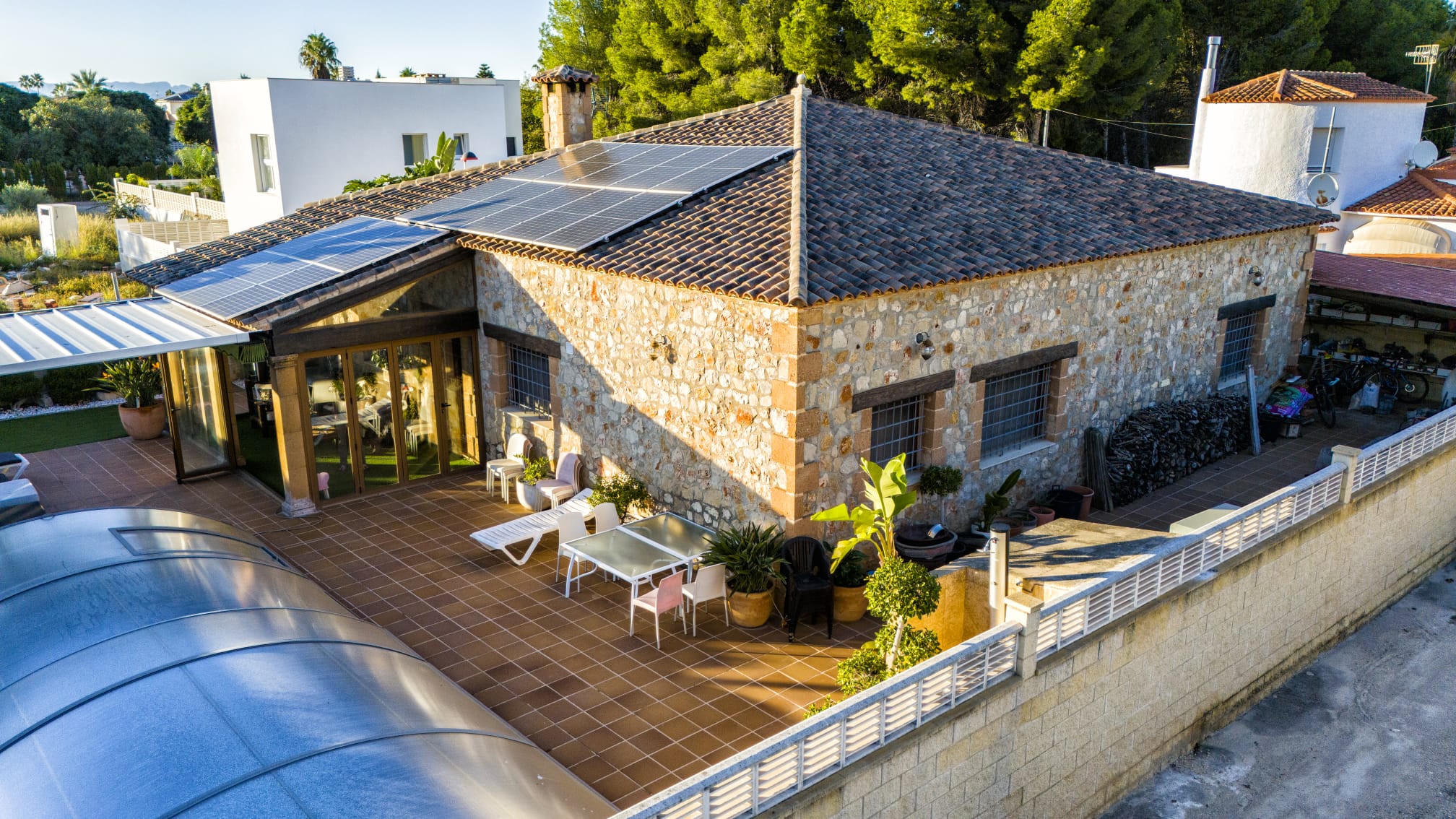 Außenansicht einer Steinvilla mit Solarmodulen, großzügiger Fliesenterrasse und einem Pool mit gebogener Schiebeüberdachung.