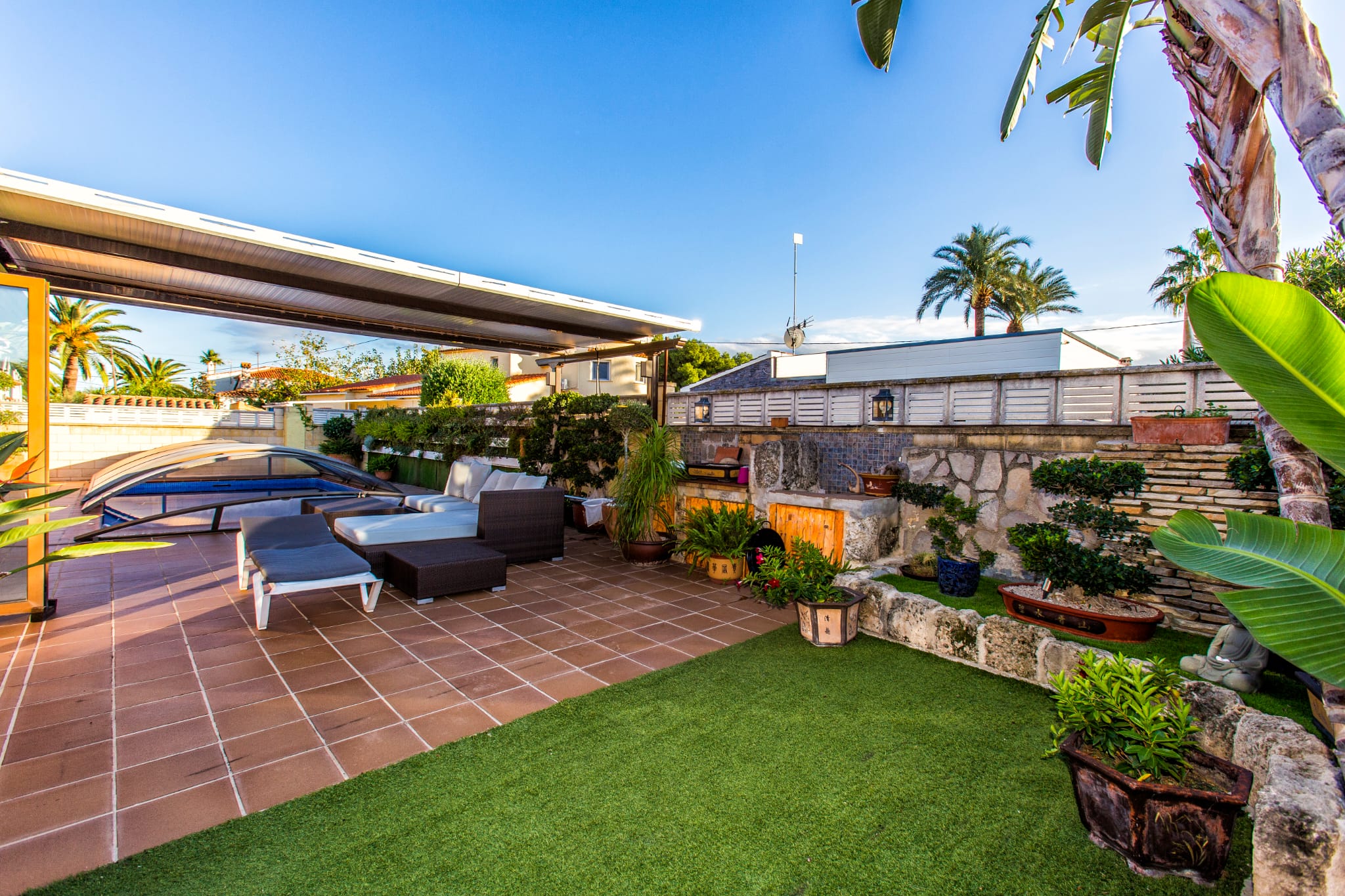 Außenterrasse mit Terrakottafliesen, Kunstrasen und Loungebereich unter einer Pergola, neben einem Pool mit Teleskopabdeckung und gemauerten Steinbeeten.