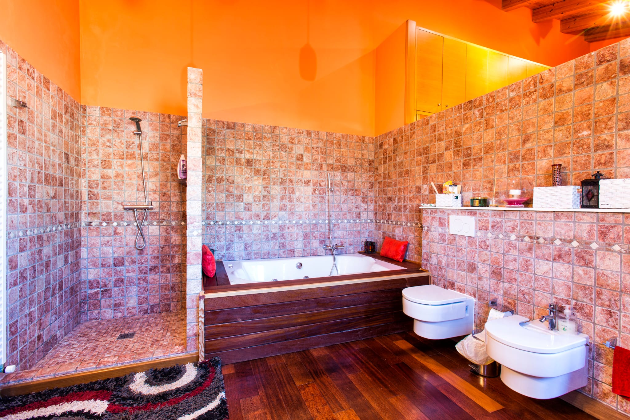 Badezimmer mit ebenerdiger Steindusche, holzverkleideter Badewanne und wandhängenden Sanitärobjekten. Orangefarbene Wände treffen auf dunkles Echtholzparkett.