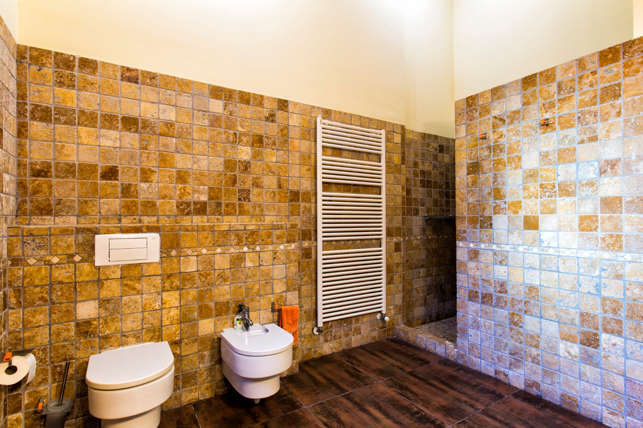 Badezimmer mit erdfarbenen Natursteinmosaik-Wänden, wandhängendem WC und Bidet, weißem Handtuchheizkörper und dunklem Boden in Holzoptik.