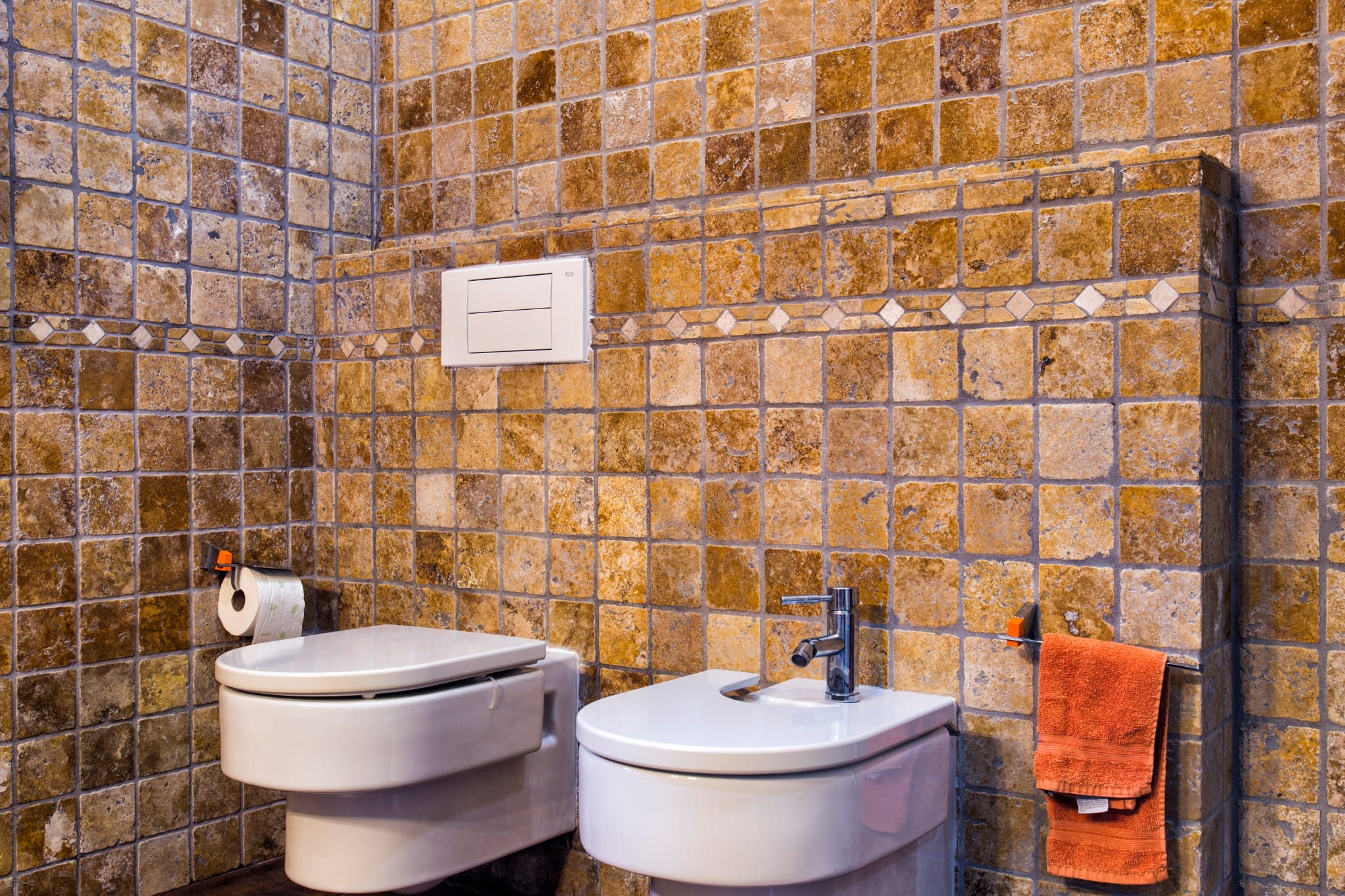 Badezimmer mit rustikalen Fliesen in Erdtönen, wandhängendem WC und Bidet sowie einer dekorativen Mosaikbordüre.