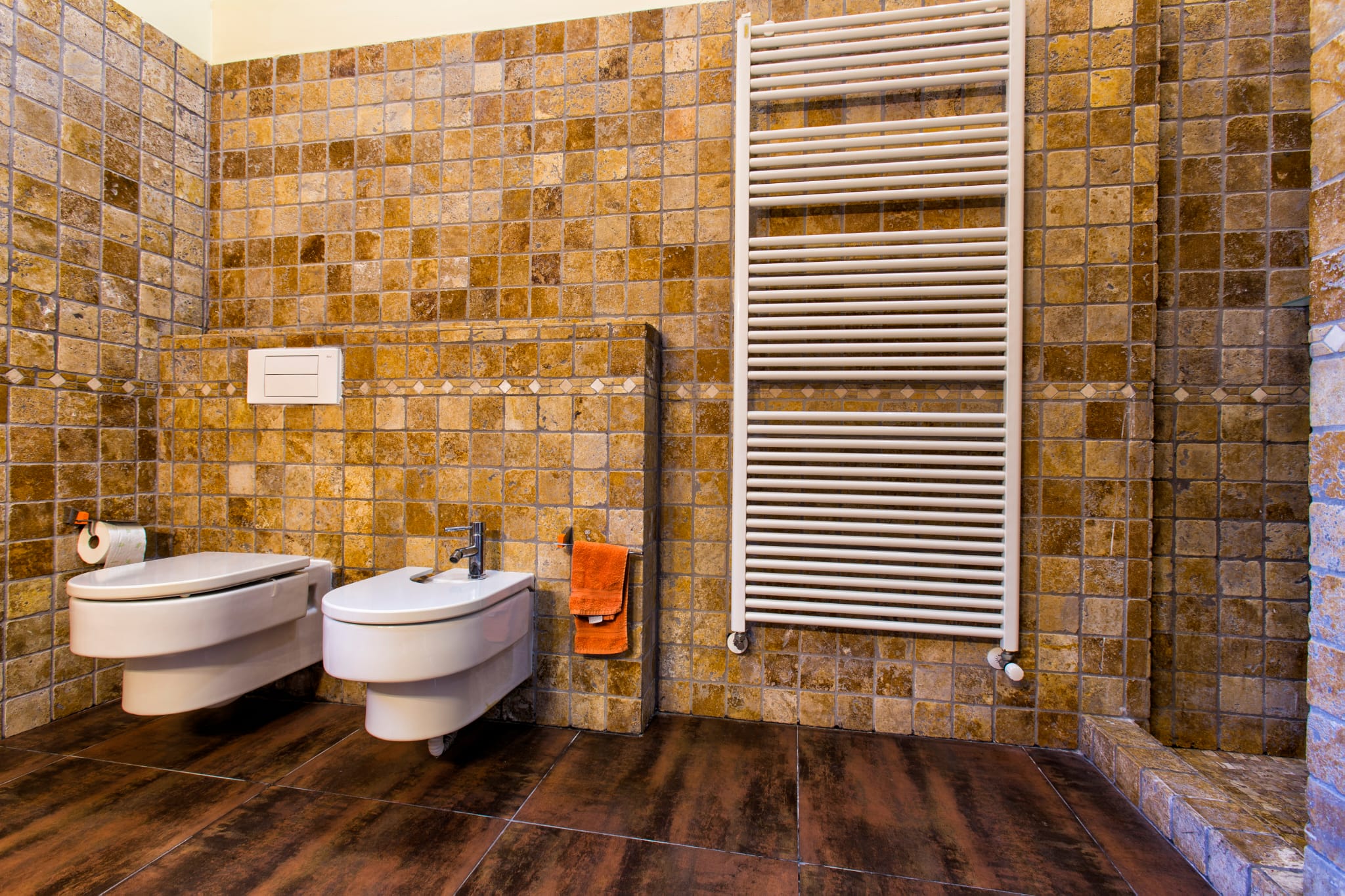 Badezimmer mit wandhängendem WC und Bidet, erdfarbenen Mosaikfliesen und einem großen weißen Handtuchheizkörper auf dunklem Fliesenboden.