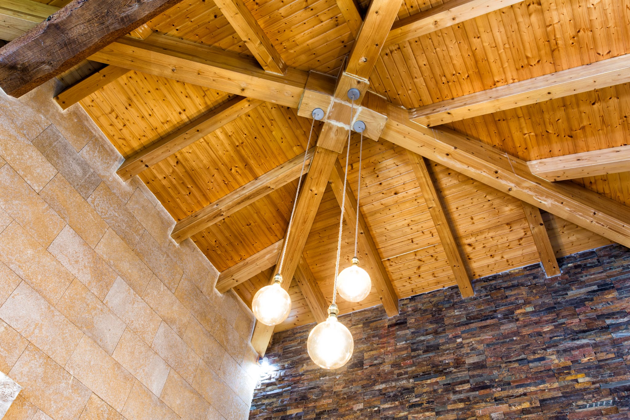 Detailansicht einer Holzbalkendecke in Sternform mit drei dekorativen Pendelleuchten vor Wänden aus Naturstein und dunklem Schiefer.