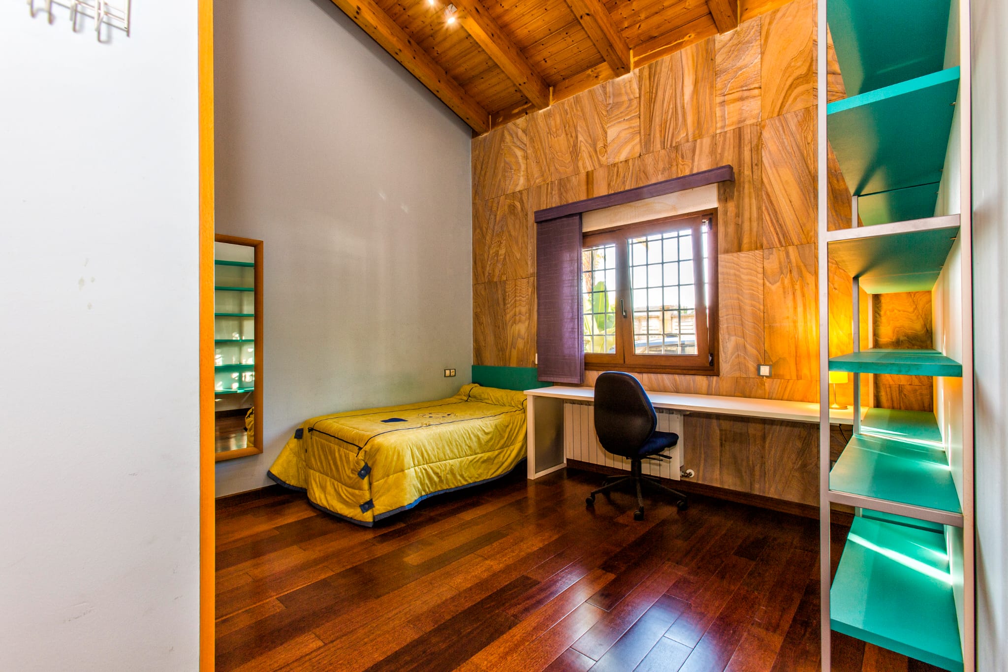 Einzelzimmer mit hohen Holzdecken, einer Akzentwand in Natursteinoptik, integriertem Arbeitsbereich und dunklem Parkettboden.
