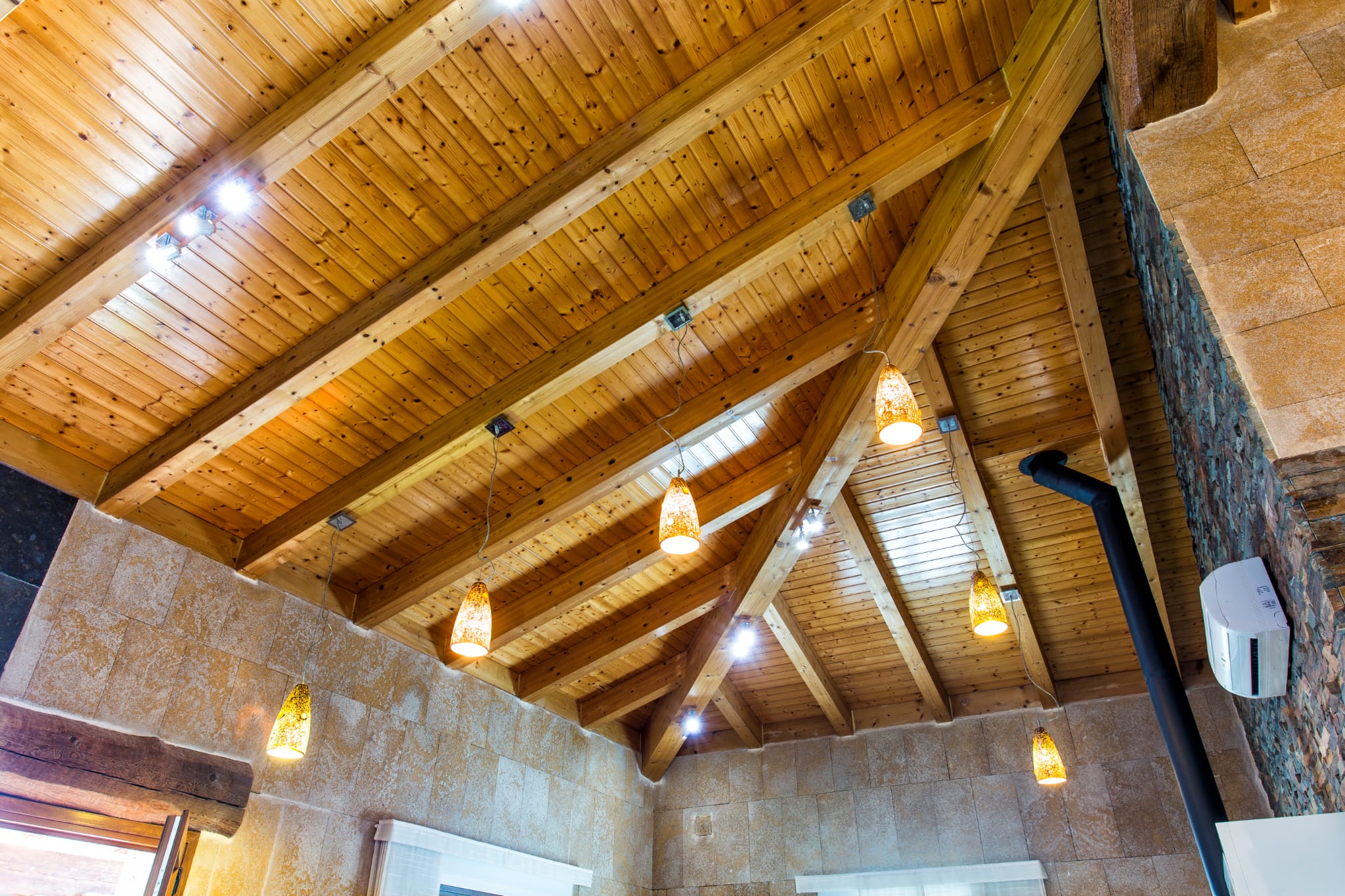 Hohe Decke mit sichtbaren Holzbalken und Kiefernholzpaneelen, kombiniert mit bernsteinfarbenen Pendelleuchten und Wänden aus Naturstein.