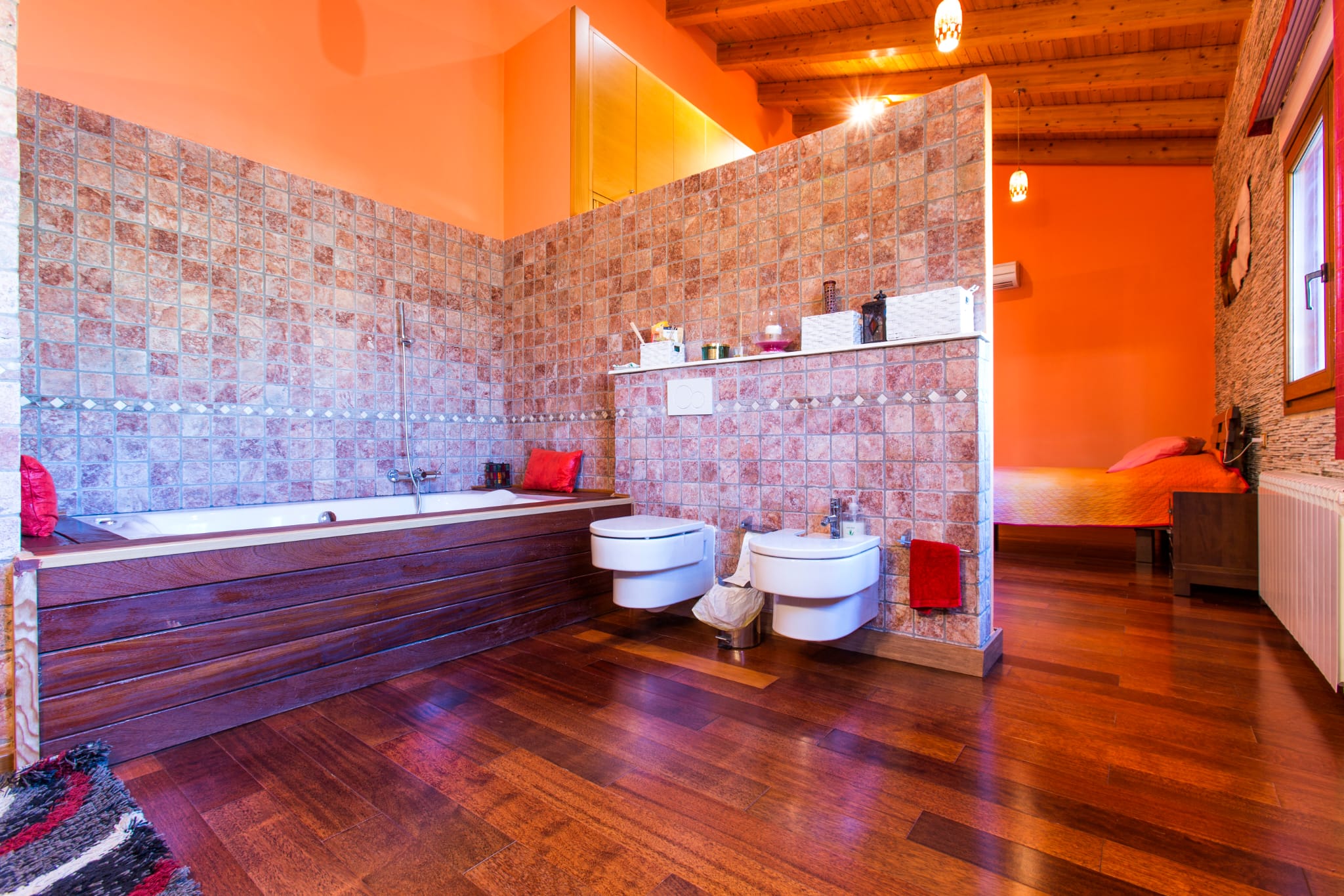 Offenes Badezimmer mit holzverkleideter Badewanne, Mosaikfliesen und wandhängenden Sanitärobjekten, integriert in ein Schlafzimmer mit Sichtbalkendecke.
