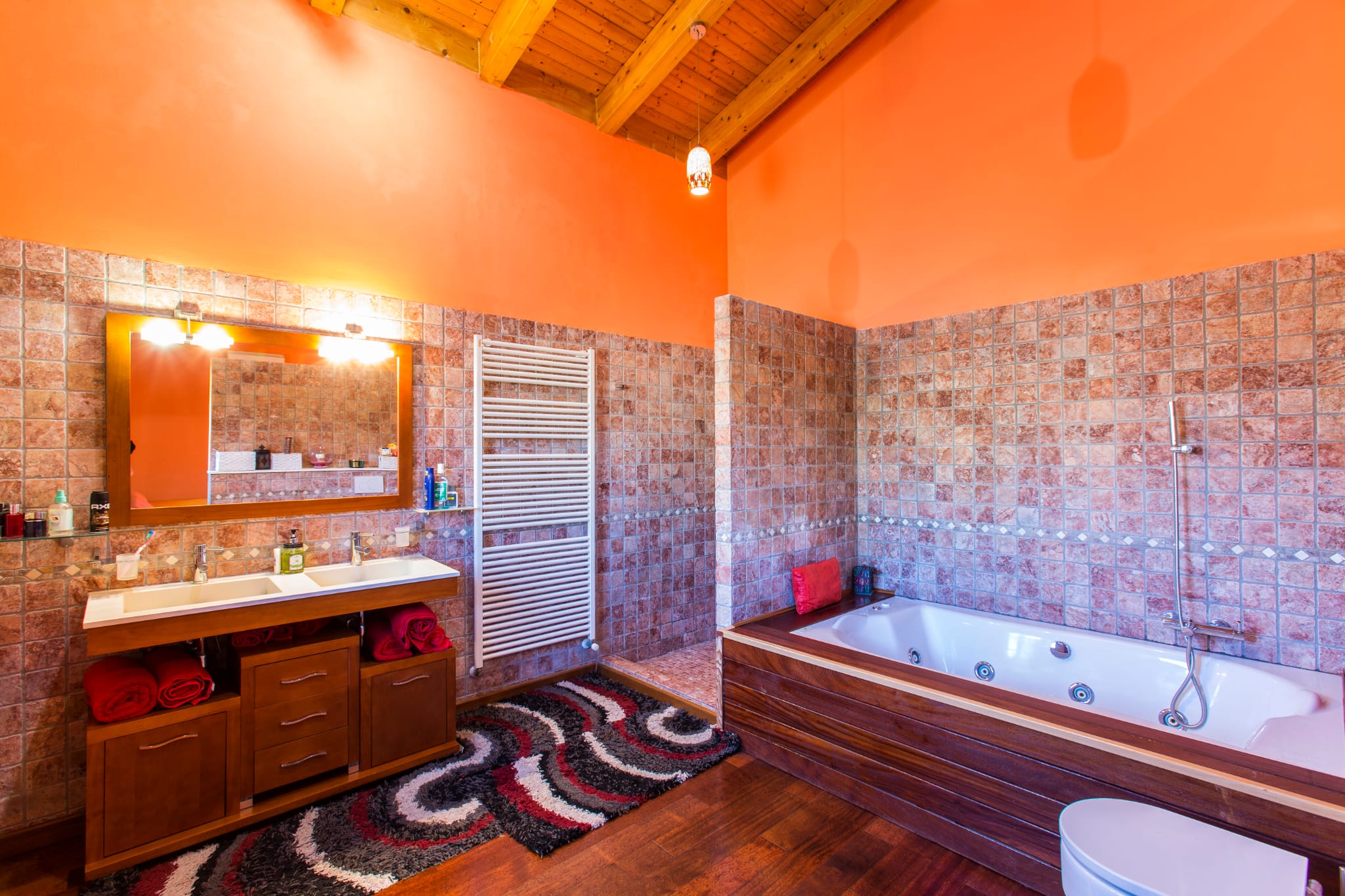 Rustikales Badezimmer mit Holzbalkendecke, Doppelwaschtisch aus dunklem Holz, Whirlpool-Badewanne und Wänden in warmen Terrakotta- und Orangetönen.