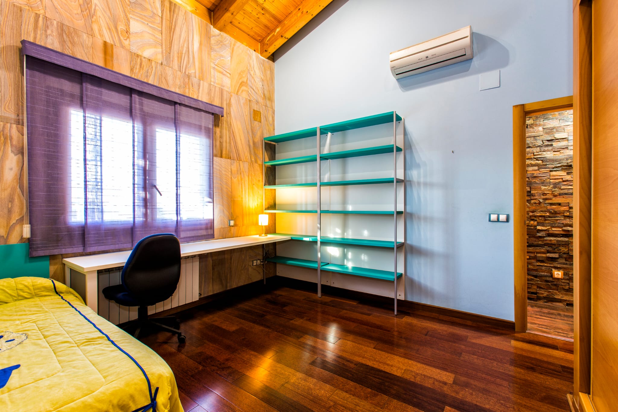 Schlafzimmer mit Arbeitsbereich, schrägen Holzdecken, türkisfarbenem Regal und weißem Schreibtisch unter einem Fenster mit lila Jalousien.