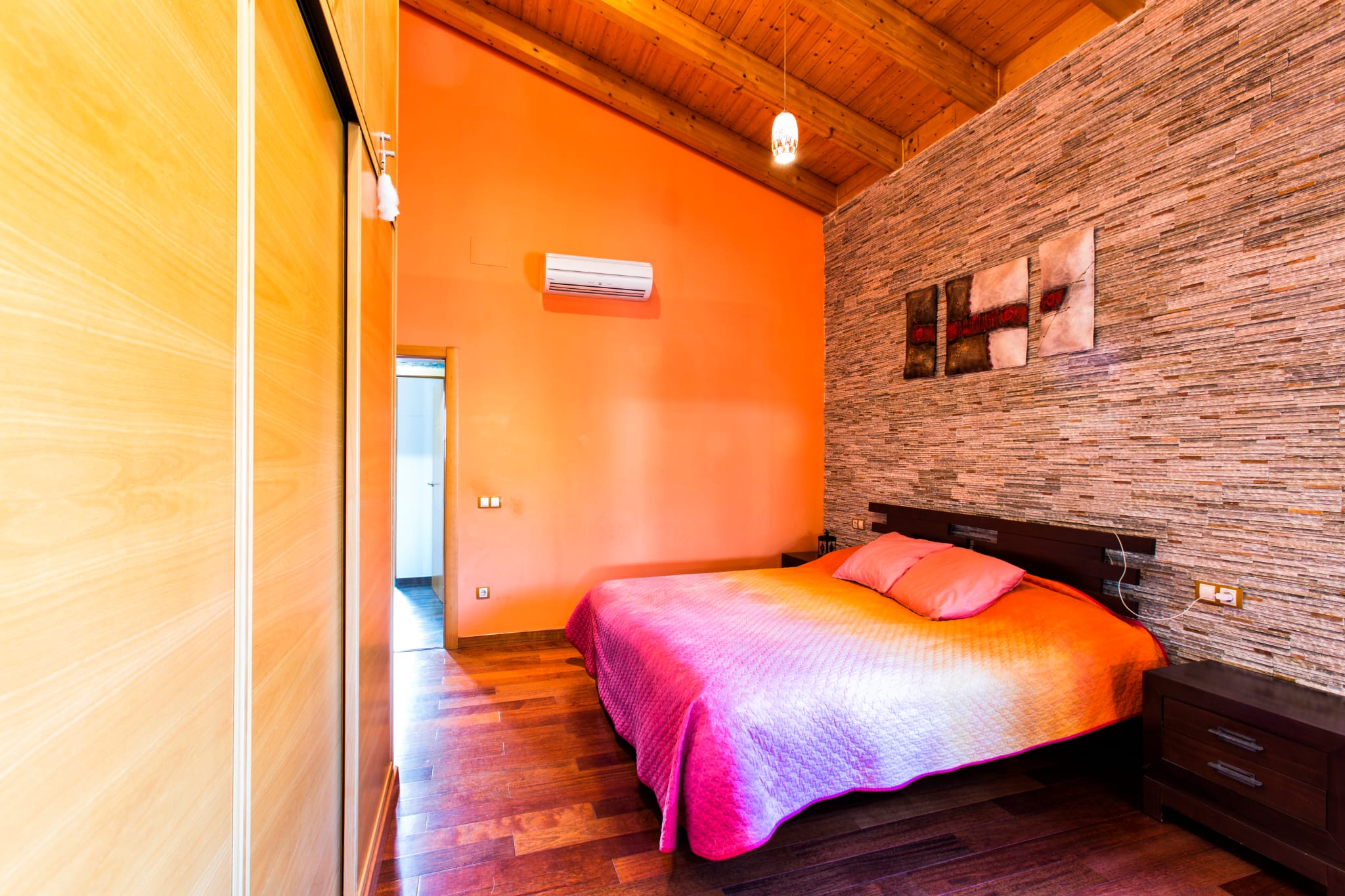 Schlafzimmer mit Dachschräge aus Holz, einer Natursteinwand und integrierten Wandschränken. Der Raum ist mit Parkettboden, Klimaanlage und dunklen Holzmöbeln ausgestattet.