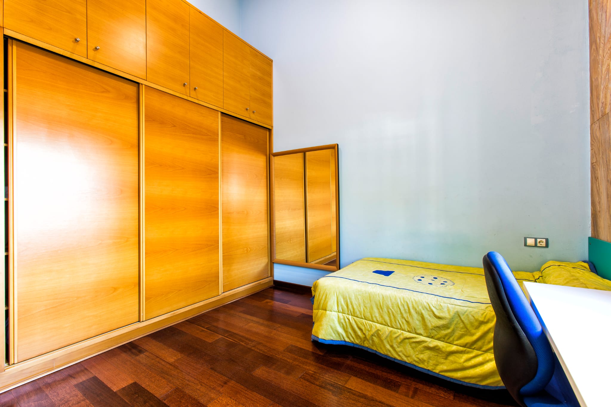 Schlafzimmer mit großem Einbauschrank aus Holz, Schiebetüren, dunklem Parkettboden und einem Einzelbett neben einem Schreibtischbereich.