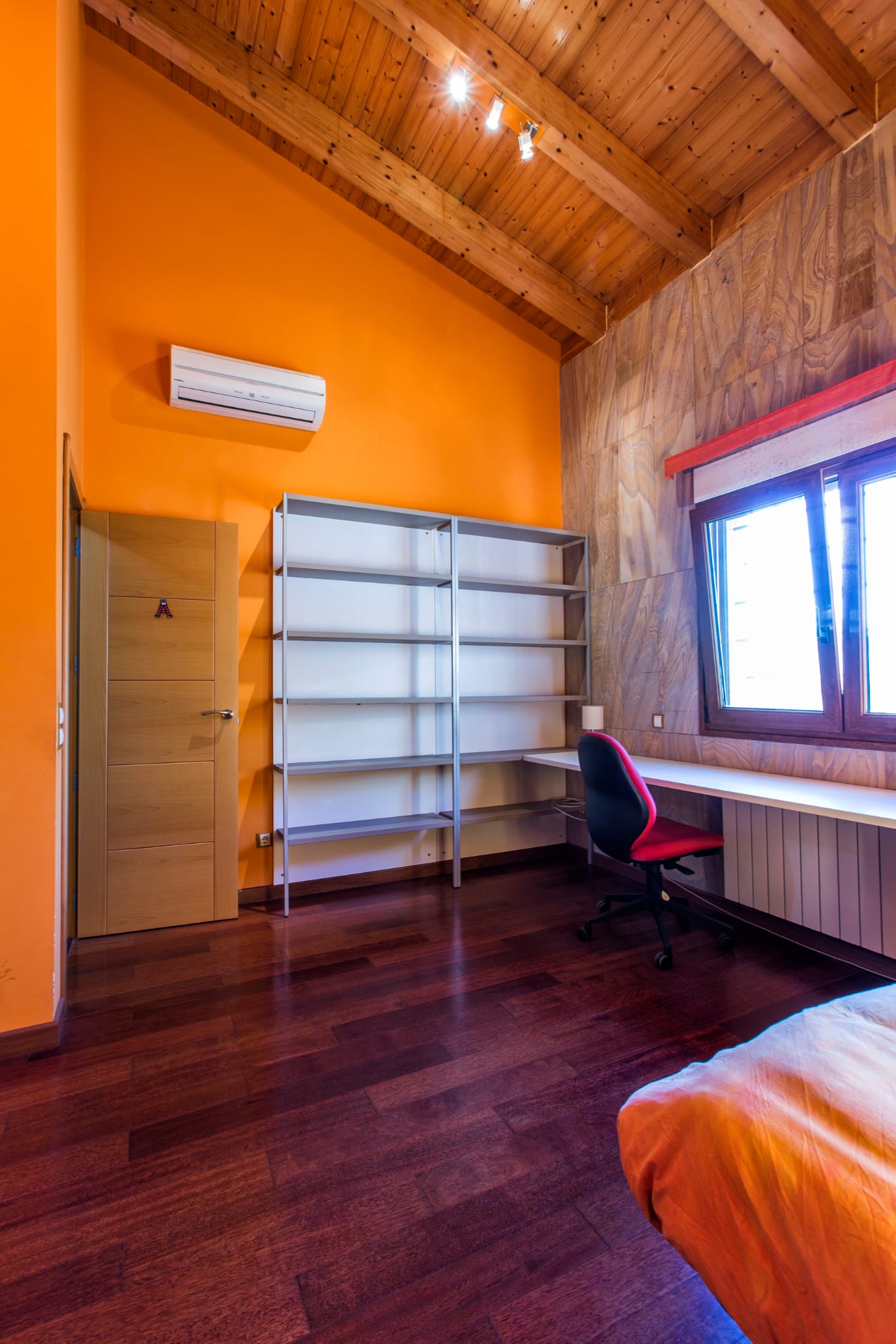Schlafzimmer mit hohen Holzdecken, orangefarbenen Wänden, integriertem Arbeitsbereich mit Schreibtisch, Metallregalen und dunklem Parkett.