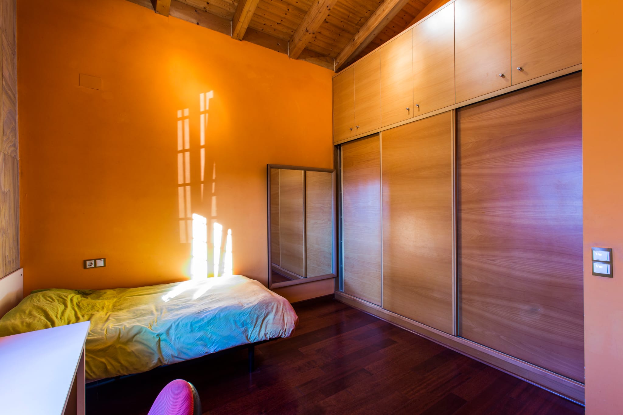 Schlafzimmer mit orangefarbenen Wänden, Sichtbalkendecke aus Holz und einem großen Einbauschrank aus Holz mit Schiebetüren.