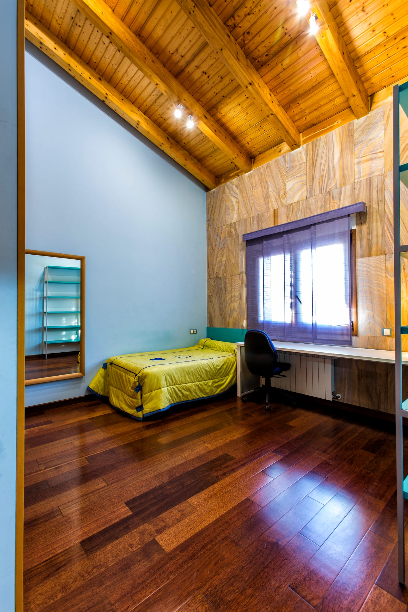 Schlafzimmer mit sichtbaren Holzbalkendecken, dunklem Parkett, integriertem Schreibtisch und einer Akzentwand aus strukturierten Paneelen.