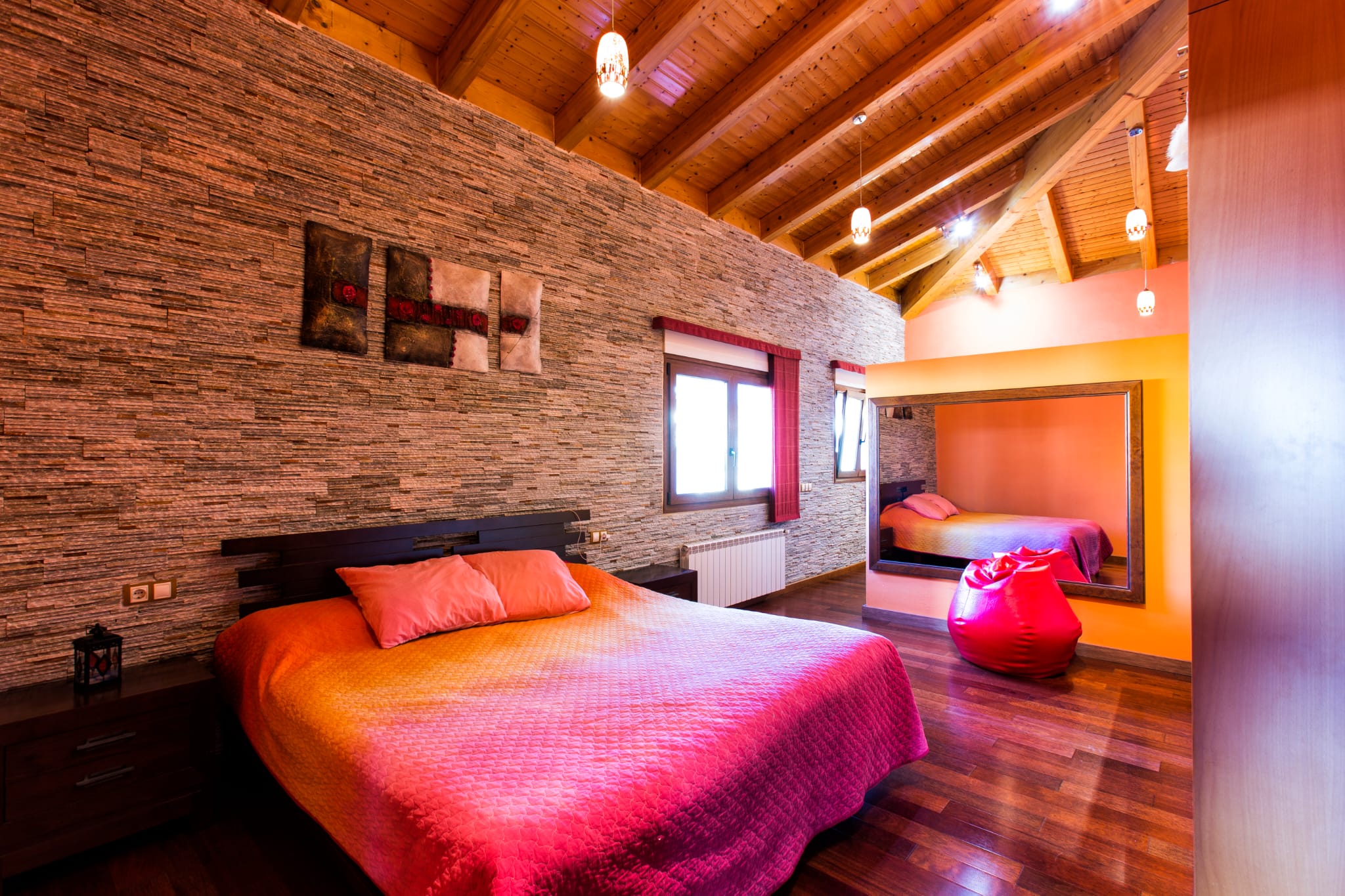 Schlafzimmer mit strukturierter Natursteinwand, Sichtbalkendecke aus Holz und dunklem Parkettboden. Ausgestattet mit einem großen Wandspiegel und Pendelleuchten.