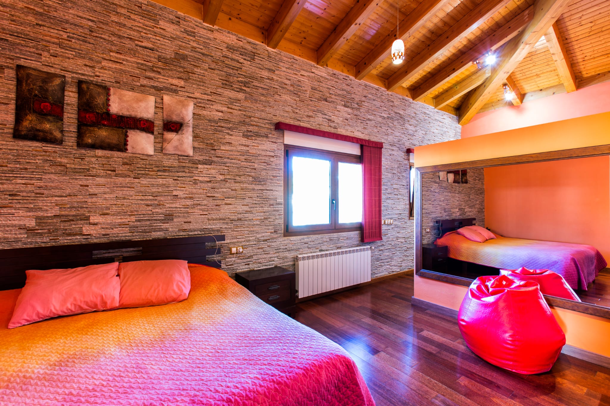 Schlafzimmer mit strukturierter Steinwand, gewölbter Holzbalkendecke und dunklem Parkett. Ein großer Wandspiegel reflektiert das Bett mit farbenfroher Bettwäsche.
