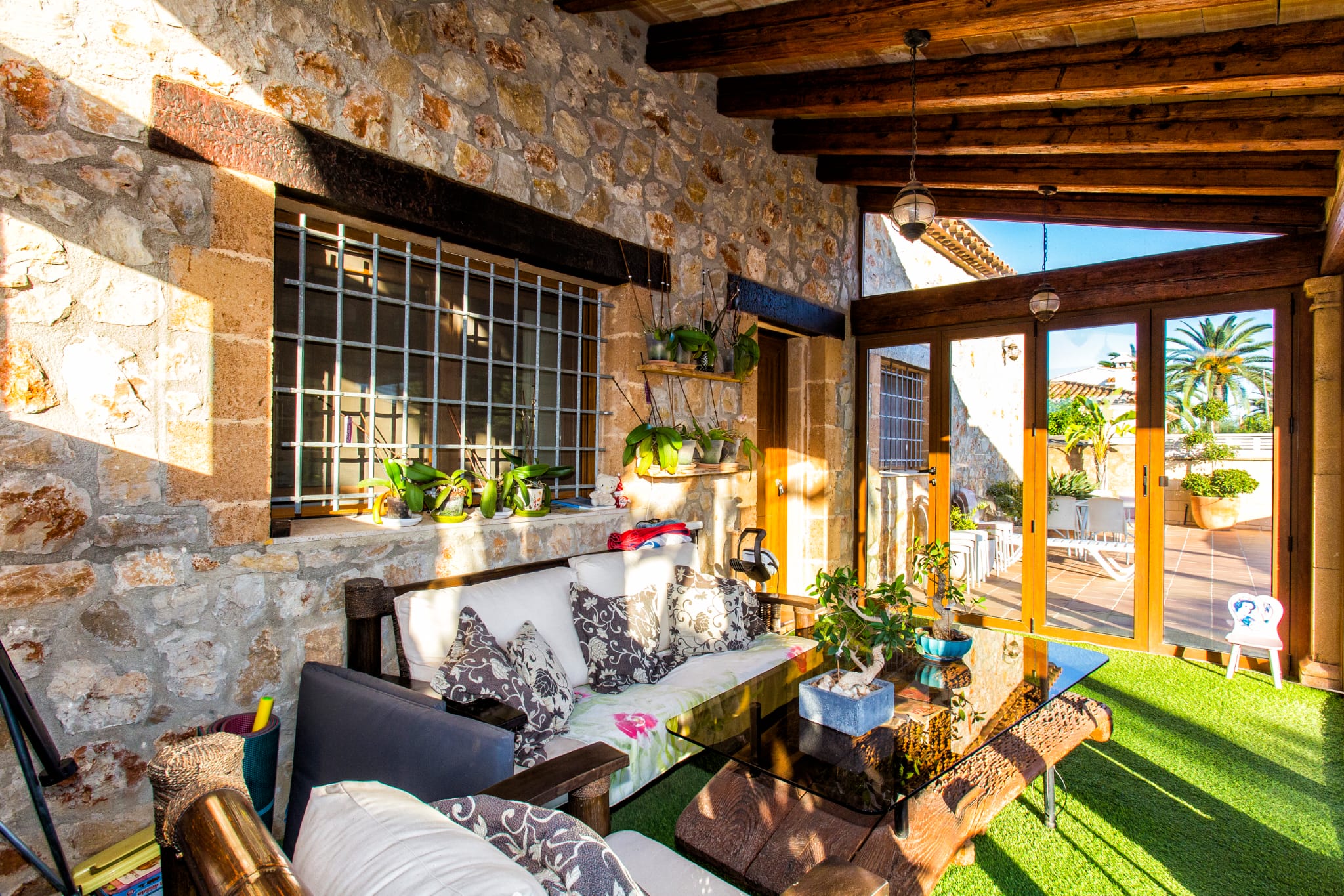 Überdachte Terrasse mit Natursteinwänden, dunklen Holzbalken und Sitzbereich mit Glastisch auf Kunstrasen, der zu einem sonnigen Innenhof führt.
