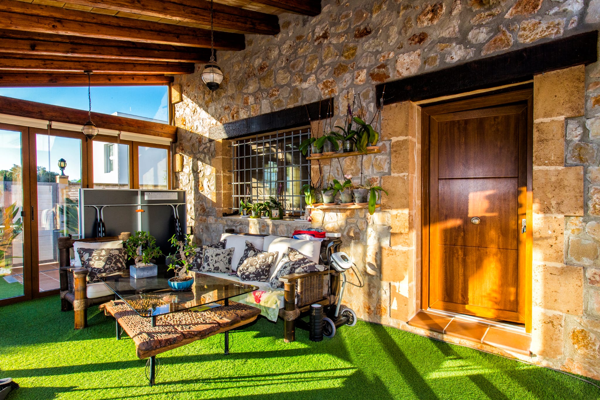 Überdachte Terrasse mit Natursteinwänden, Holzbalkendecke und Kunstrasen. Ausgestattet mit Loungebereich, Treibholz-Couchtisch und Orchideenregalen.