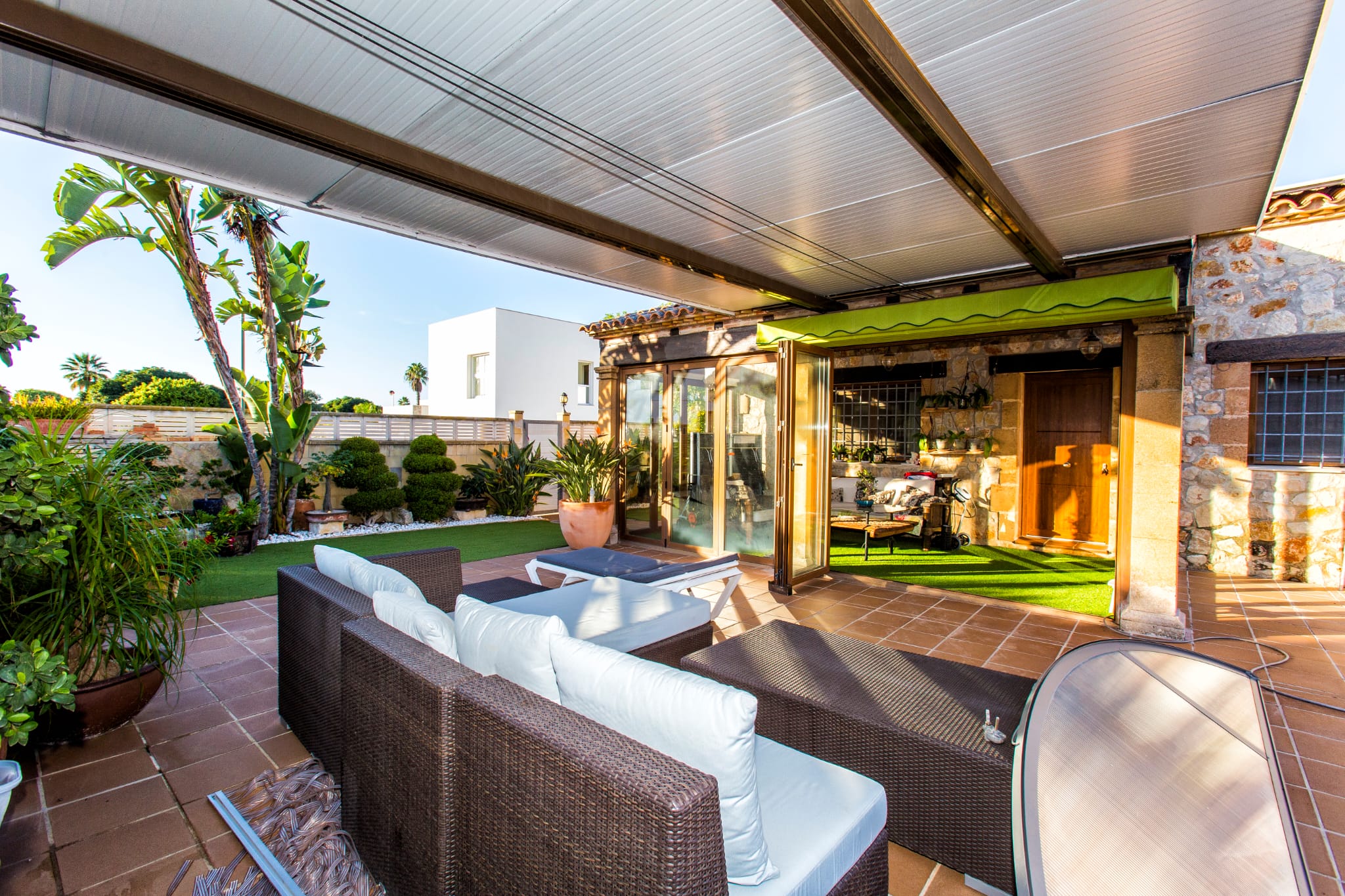 Überdachte Terrasse mit Polyrattan-Sitzgruppe, Terrakotta-Bodenfliesen und gepflegtem Garten mit Kunstrasen und tropischen Pflanzen vor Steinfassade.