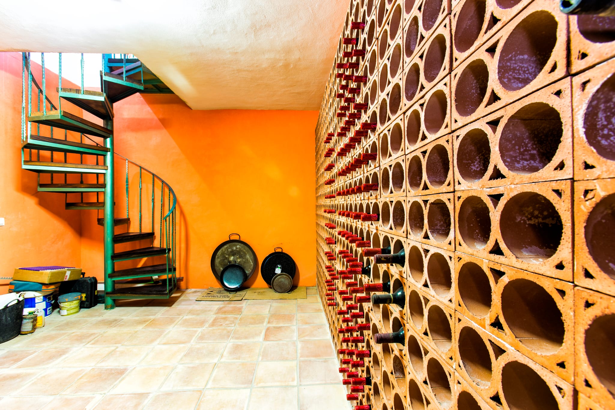 Weinkeller mit Flaschenregal aus Terrakotta-Waben, orangefarbener Wand und einer grünen Wendeltreppe aus Metall mit dunklen Holzstufen.