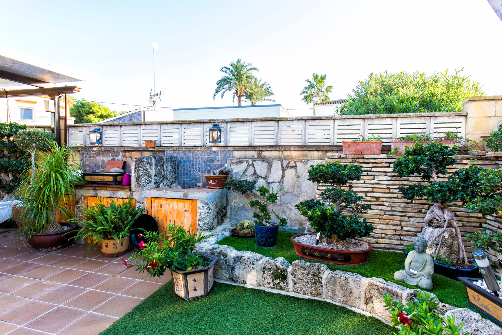 Zen-Garten auf der Terrasse mit Kunstrasen, Natursteinmauern und einer Bonsai-Sammlung. Integrierte Steintheke mit blauem Mosaik-Hintergrund.