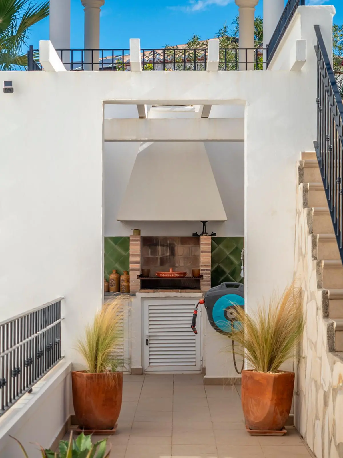 Außen-Grillbereich mit gemauertem Grill, weißer Abzugshaube und grünen Fliesen, ergänzt durch Terrakotta-Pflanzgefäße und weiße Architekturwände.