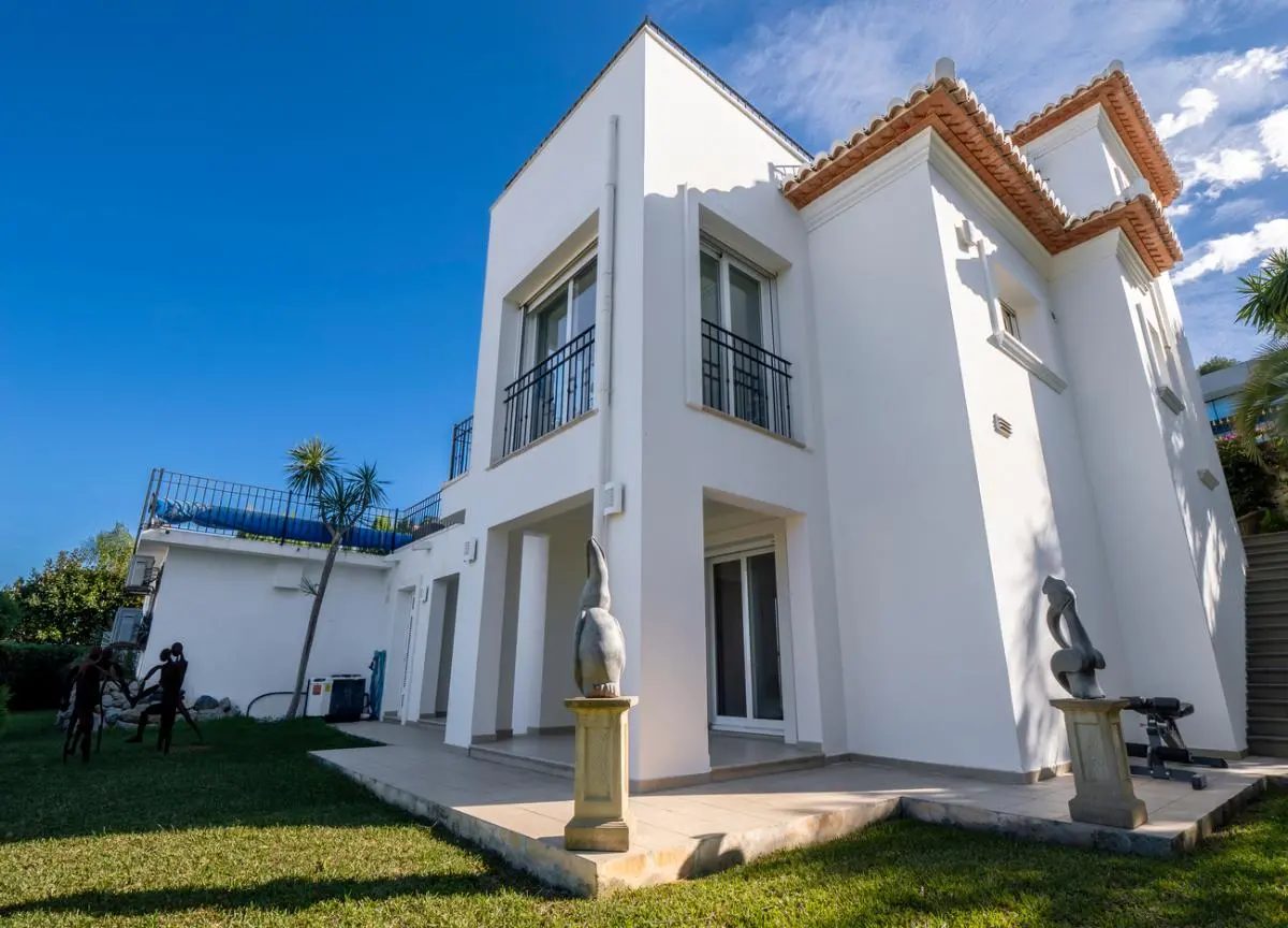 Außenansicht einer weißen Villa mit Terrakotta-Dachziegeln, schwarzen Eisenbalustraden und gepflegtem Rasen mit dekorativen Steinskulpturen auf Sockeln.