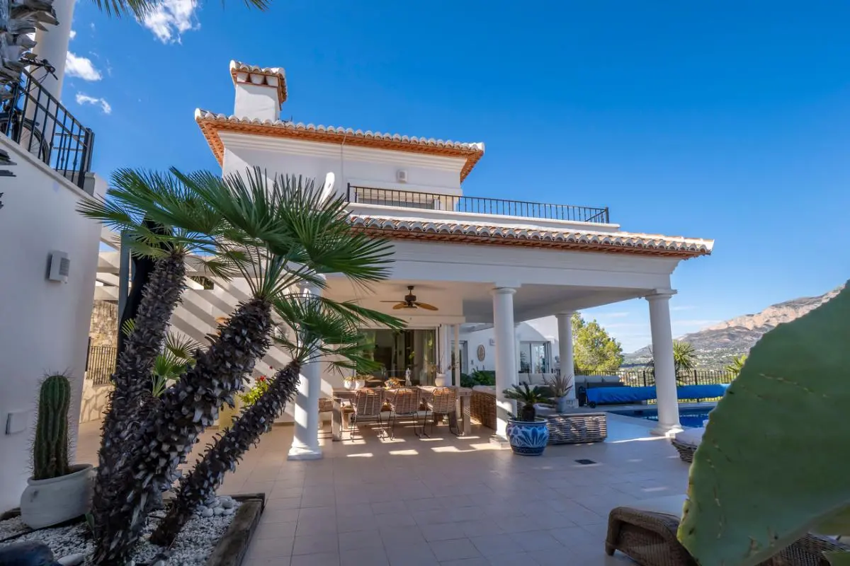 Außenansicht einer weißen Villa mit überdachter Säulenterrasse, Essbereich im Freien und mediterranem Garten mit Panoramablick auf die Berge.