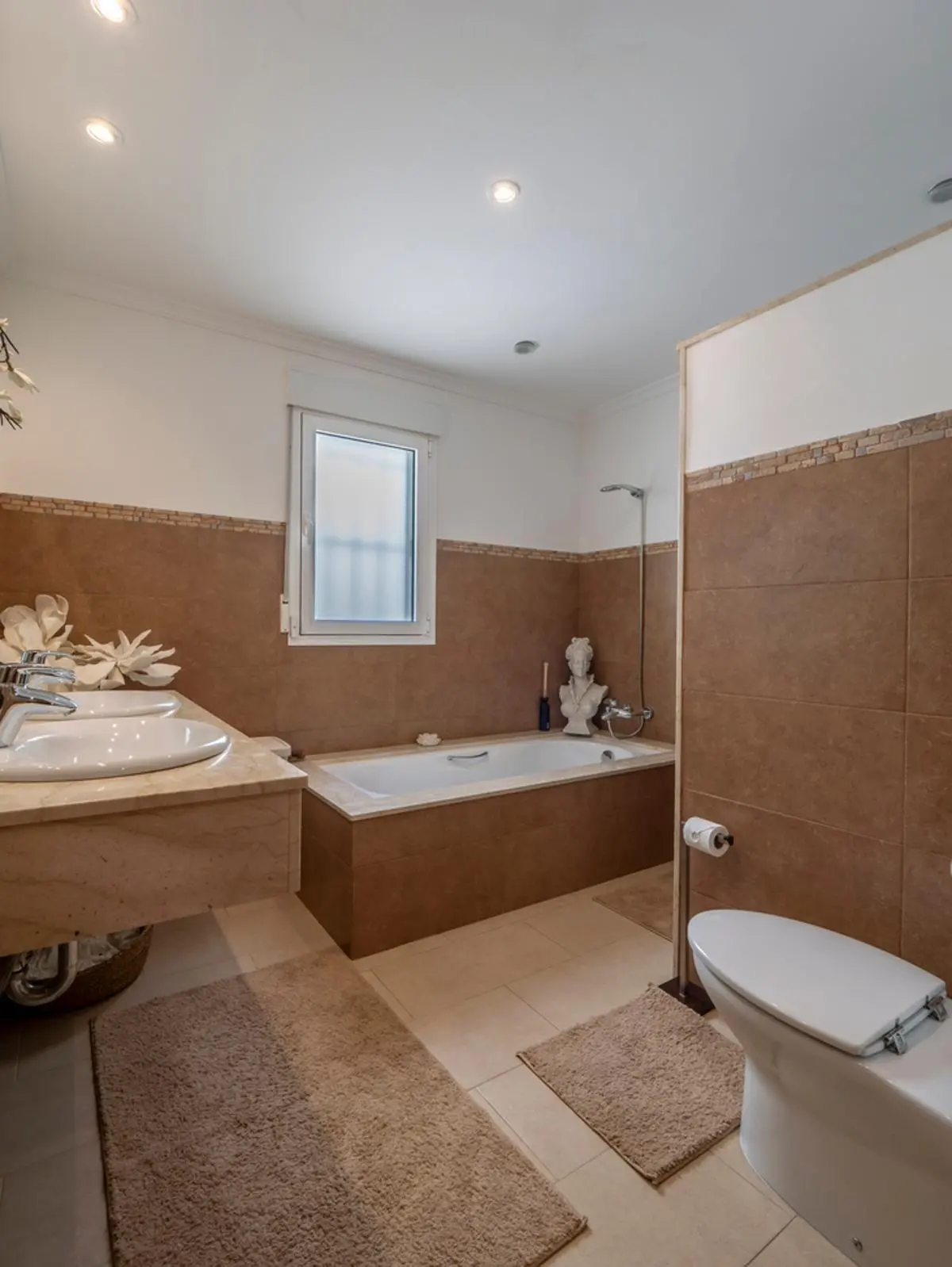 Badezimmer mit Doppelwaschbecken auf Marmorplatte, eingebauter Badewanne mit braunen Fliesen und Deckeneinbaustrahlern.