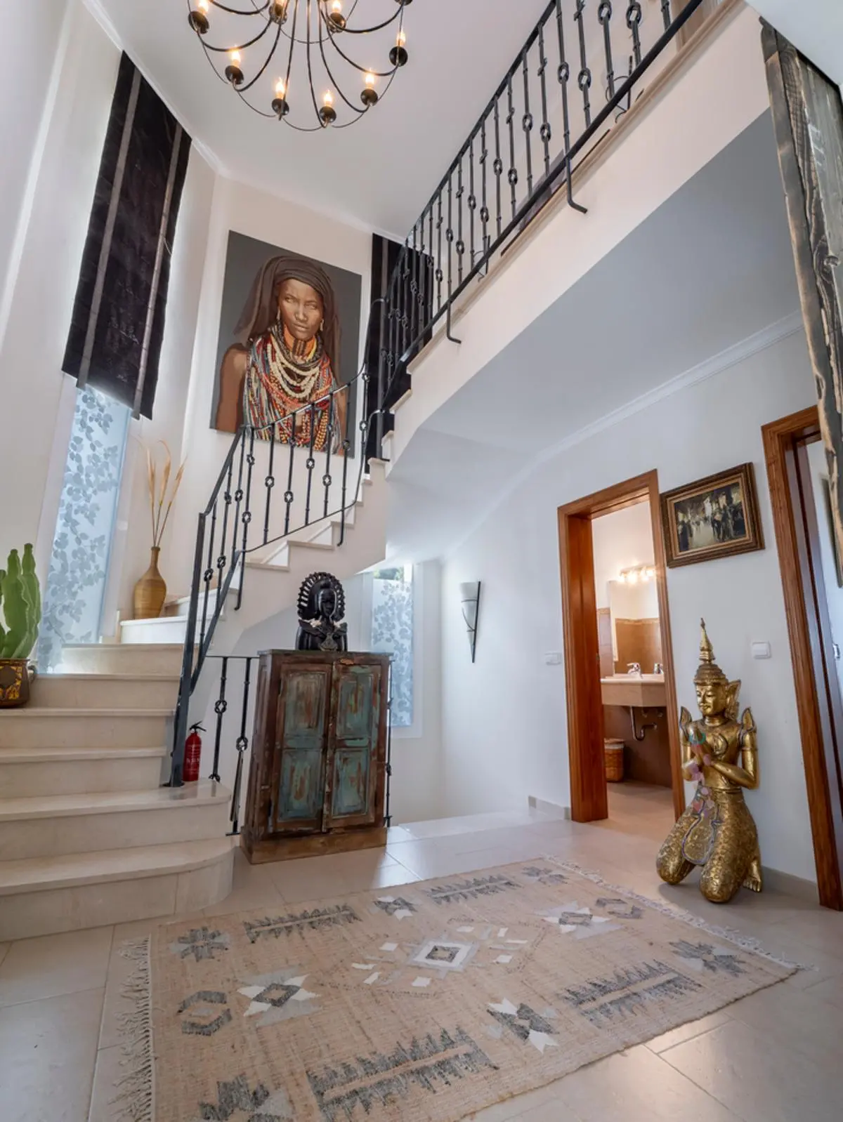 Eingangsbereich mit Marmortreppe, schmiedeeisernem Geländer, großem Porträtgemälde und eklektischem Dekor mit goldener Statue und gemustertem Teppich.
