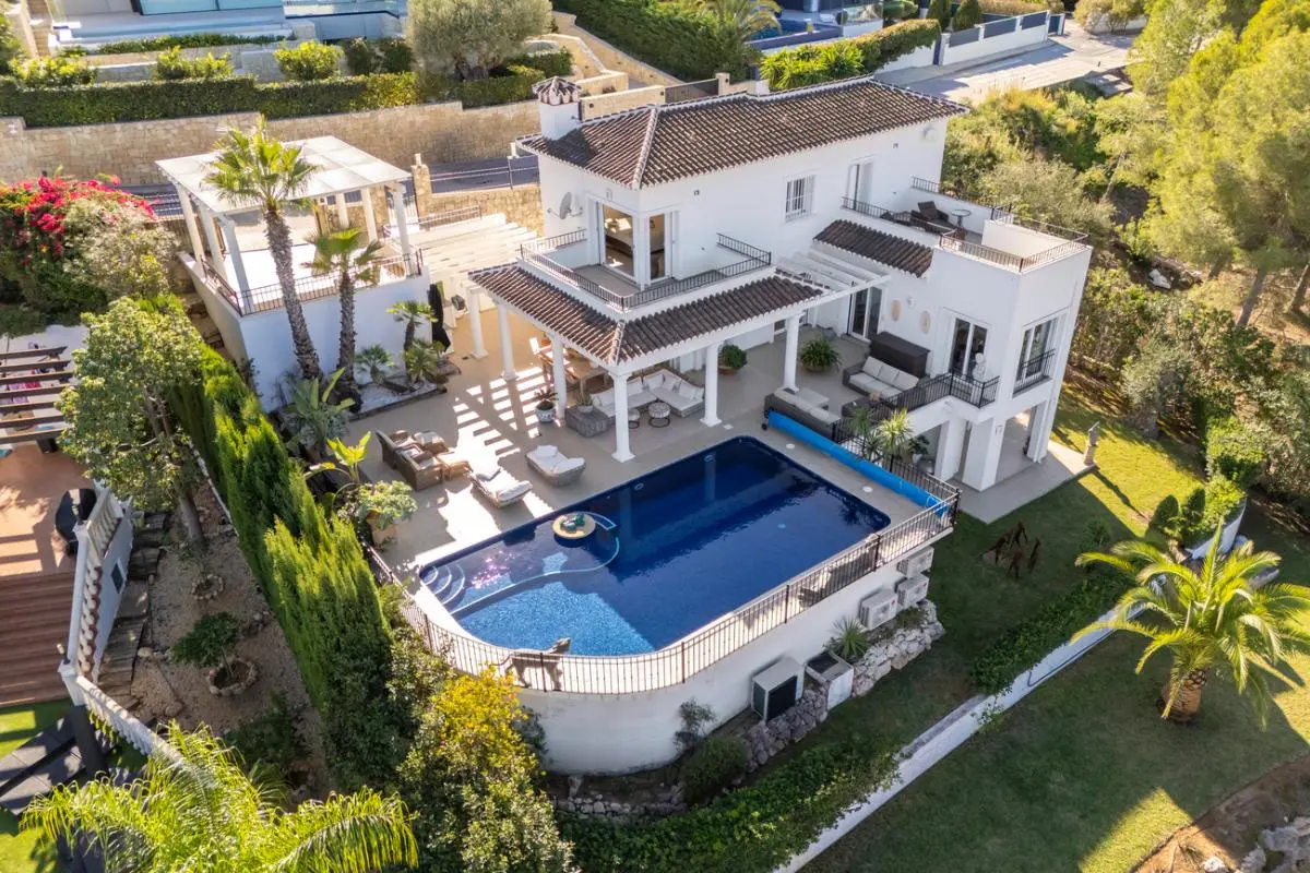 Luftaufnahme einer weißen mediterranen Villa mit großem Pool, mehreren überdachten Terrassen mit Säulen und angelegten Gärten mit Palmen.