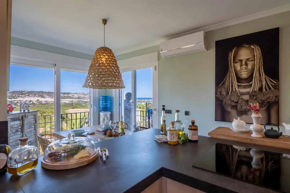 Moderne Küche mit dunkler Arbeitsplatte, Induktionskochfeld und Essbereich mit Korblampe, ergänzt durch einen Panoramablick auf die Küste durch Glastüren.