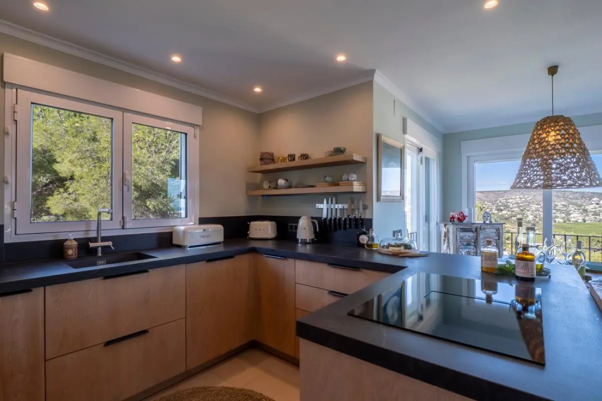 Moderne Küche mit hellen Holzschränken, dunklen Arbeitsplatten und Induktionskochfeld. Große Fenster bieten Tageslicht und Ausblick auf Pinien und das umliegende Tal.