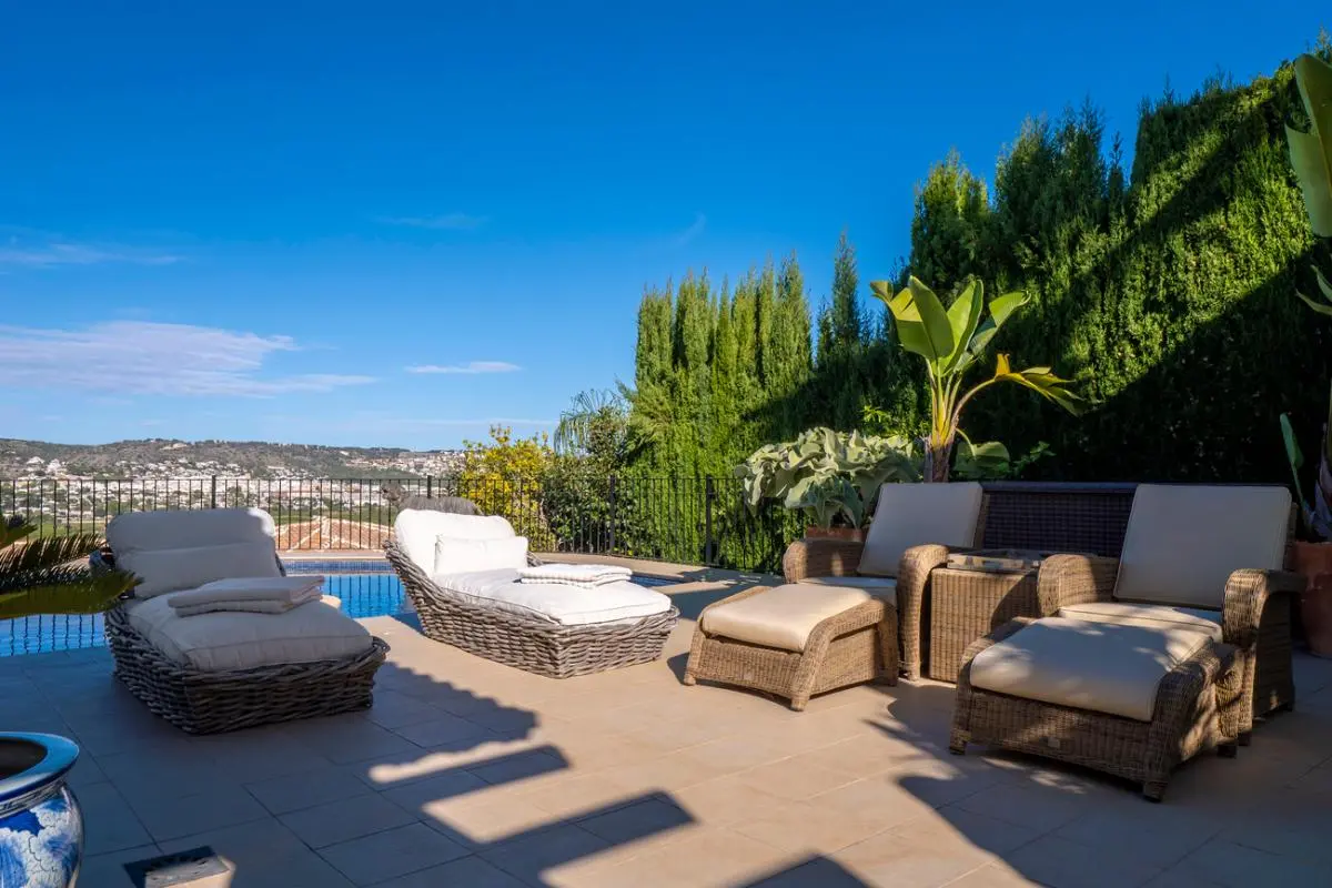 Sonnenterrasse mit Rattanchaiselongues und weißen Auflagen am Pool, mit weitem Blick auf die Hügel und dichte mediterrane Bepflanzung.