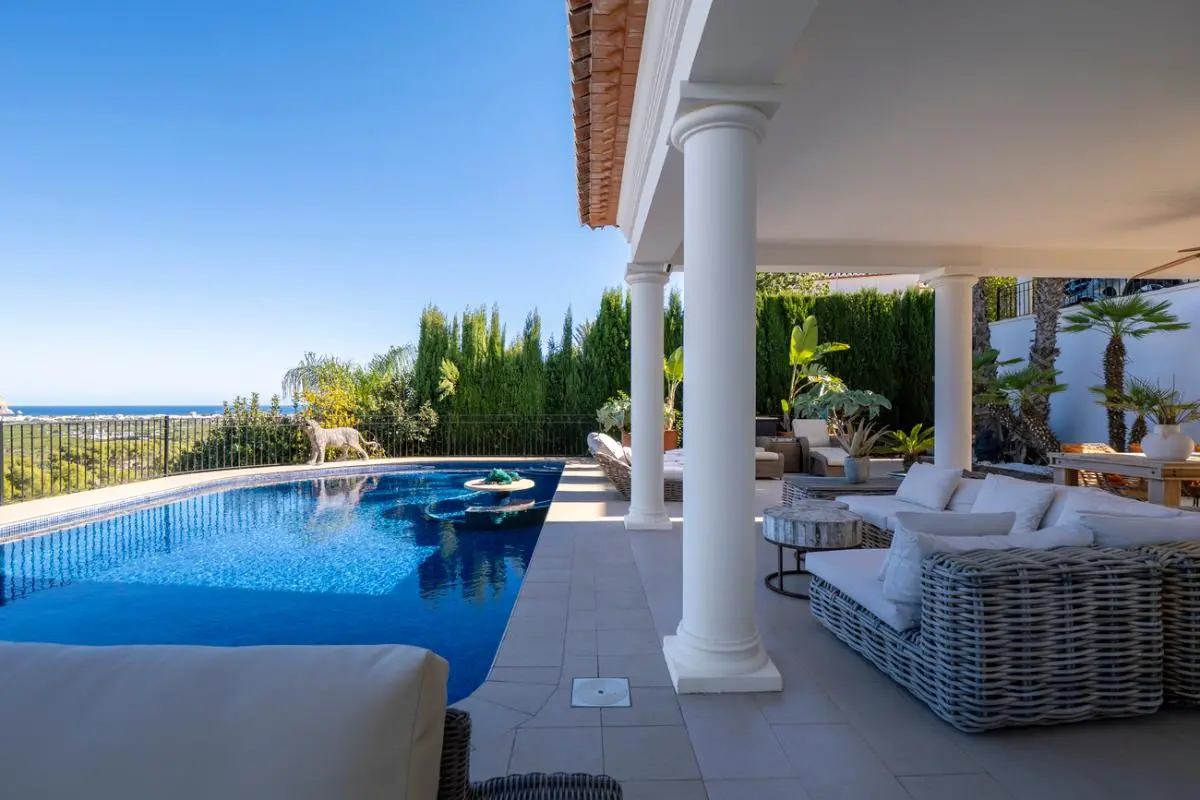 Überdachte Terrasse mit klassischen weißen Säulen und Korbmöbeln an einem Mosaikpool mit weitem Blick auf das Mittelmeer.