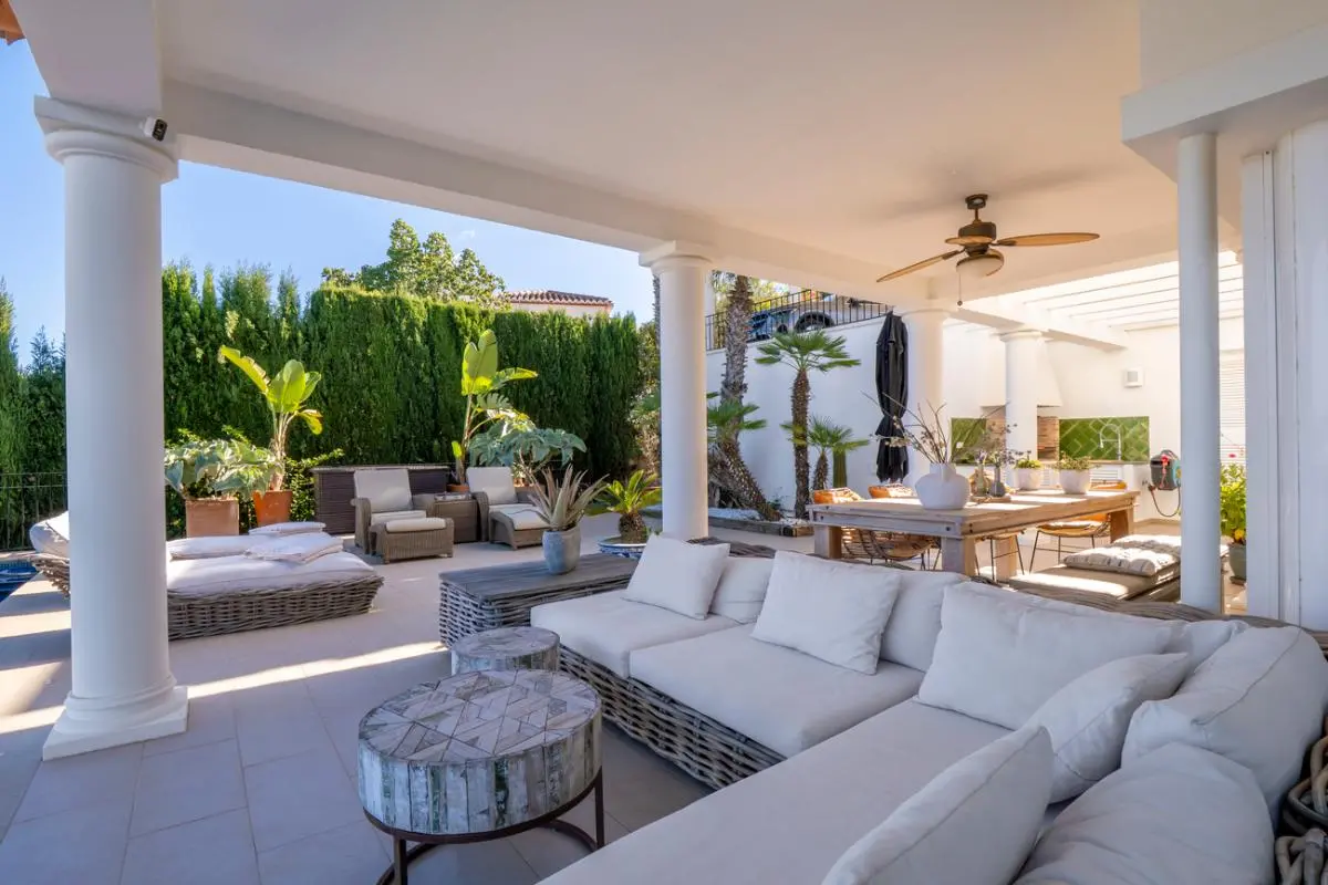 Überdachte Terrasse mit weißen Säulen, großem Rattansofa und Außen-Essbereich mit Deckenventilator, umgeben von tropischer Bepflanzung.