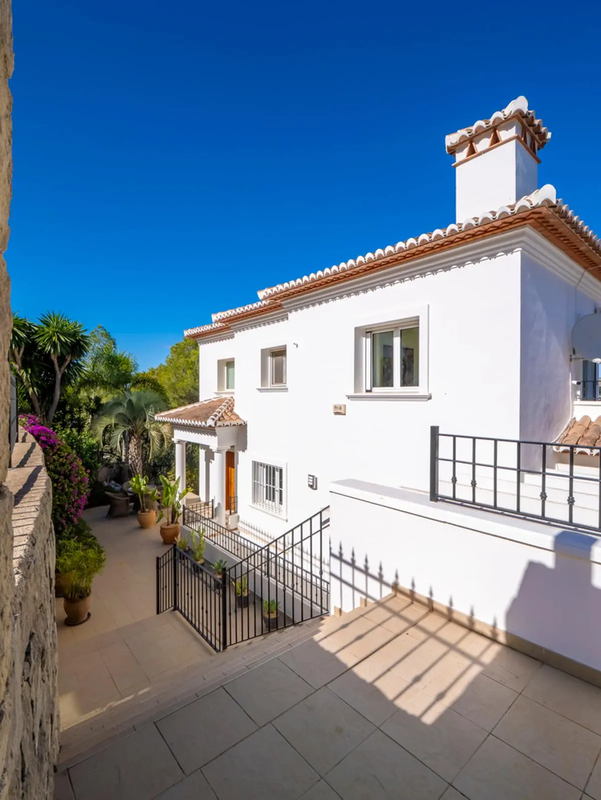 Weiße mediterrane Villa mit Terrakotta-Ziegeldach, dekorativem Schornstein und schwarzen Schmiedeeisengeländern über einer gefliesten Terrasse mit Bepflanzung.