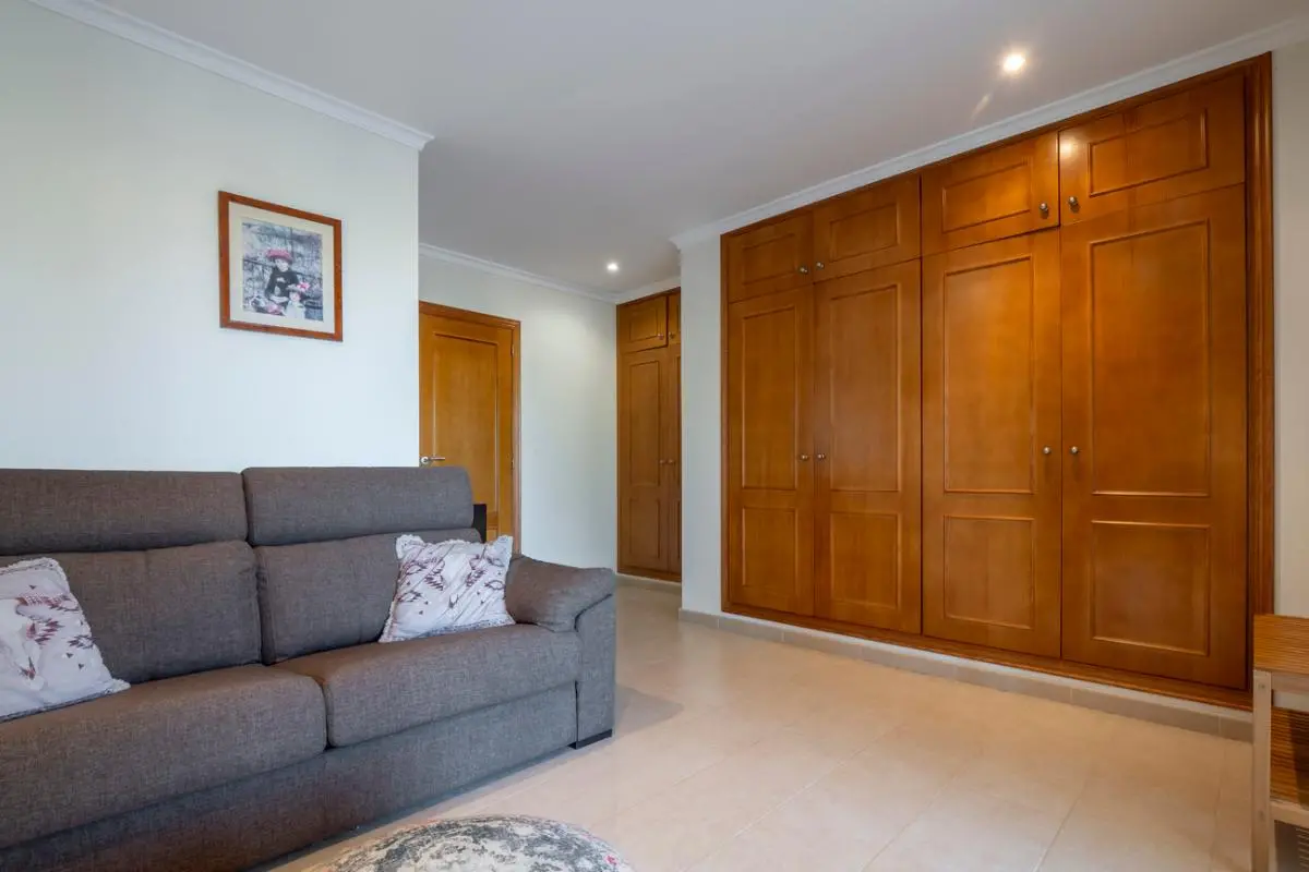 Zimmer mit einem großen Einbauschrank aus Holz, einem grauen Stoffsofa und hellen Fliesenböden unter modernen Deckeneinbaustrahlern.