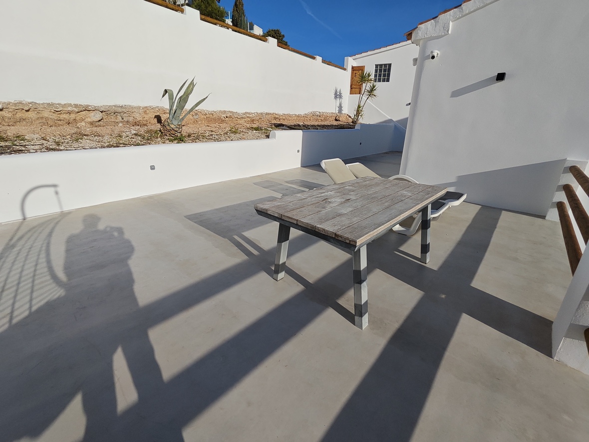 Außen-Terrasse mit Mikrozementboden, einem rustikalen Holztisch und weißen Mauern unter klarem blauem Himmel.