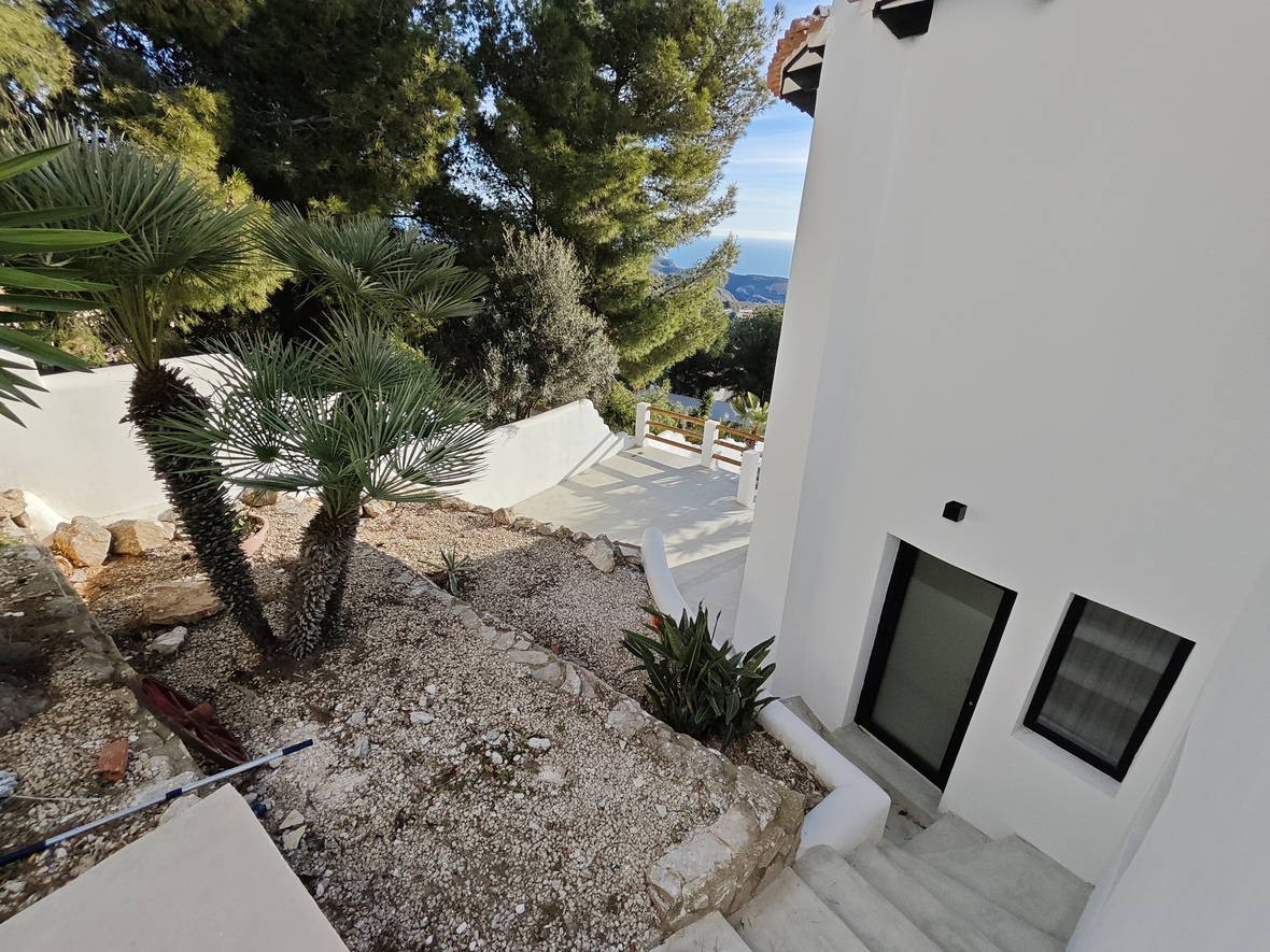 Außenansicht der weißen Villa mit Kiesgarten, Palmen und Steinstufen, die zu einem Eingang mit schwarzen Rahmen und Fernblick auf das Meer führen.