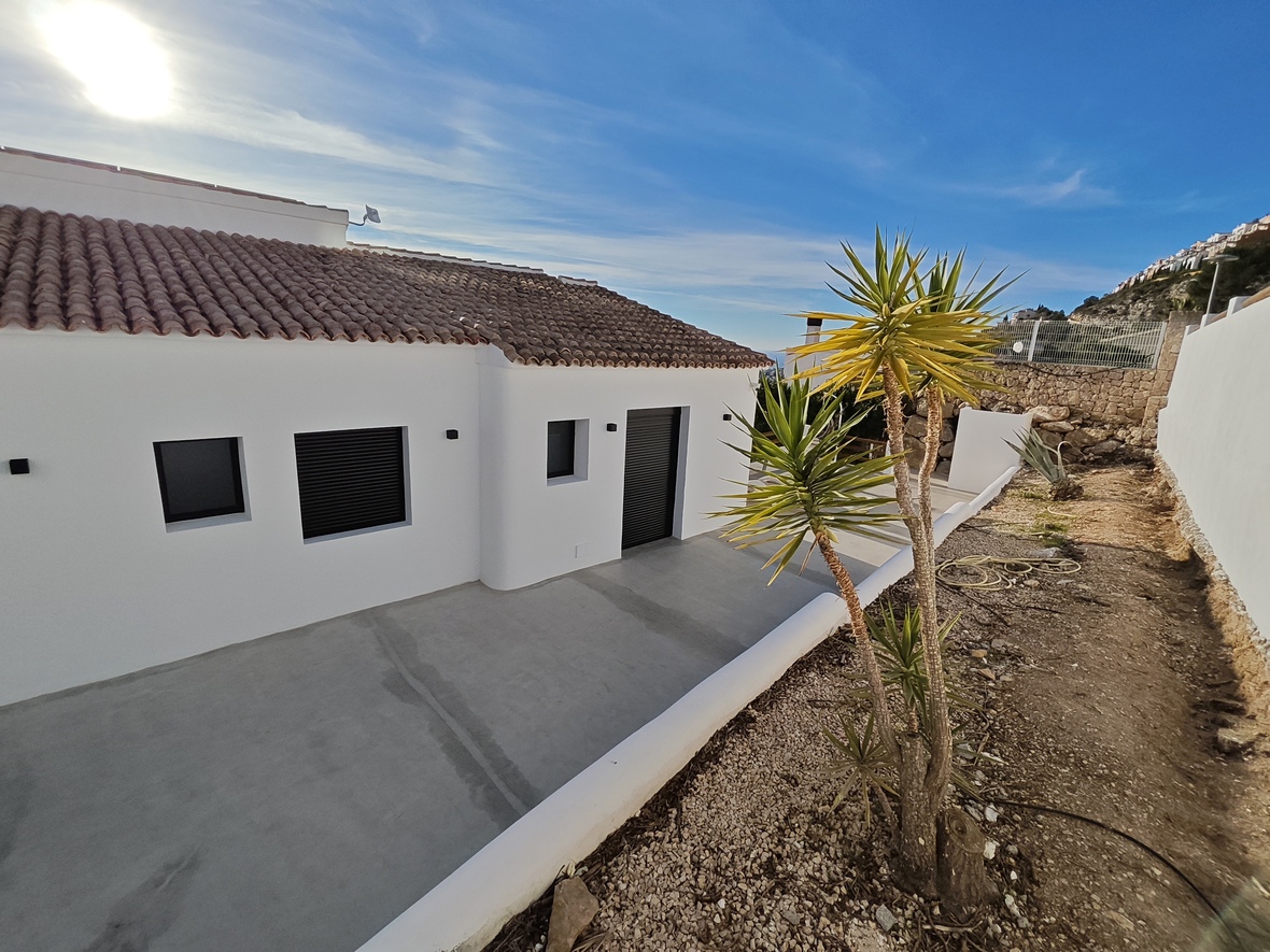 Außenansicht einer minimalistischen weißen Villa mit Ziegeldach, dunklen Rollläden und Betonterrasse neben einer Yucca-Palme und Natursteinmauer.