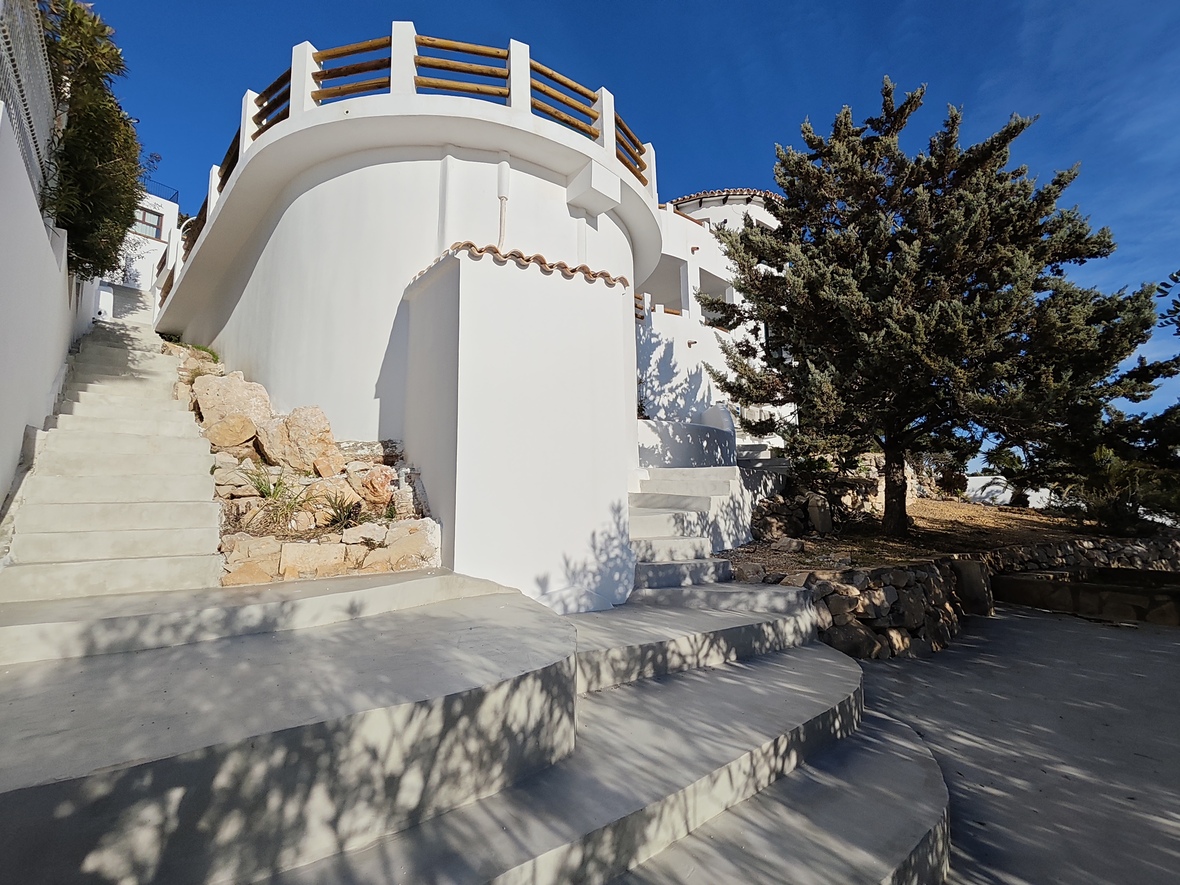 Außenansicht einer weißen mediterranen Villa mit geschwungenen Wänden, breiten Betontreppen und einer Rundterrasse mit Holzgeländer.