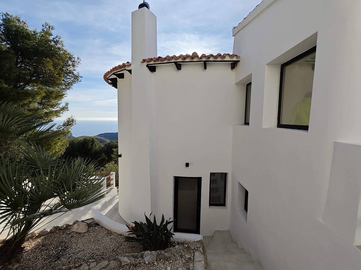 Außenansicht einer weißen mediterranen Villa mit geschwungener Fassade, Terrakotta-Ziegeldach und Treppenaufgang mit Blick auf das Meer und die Berge.