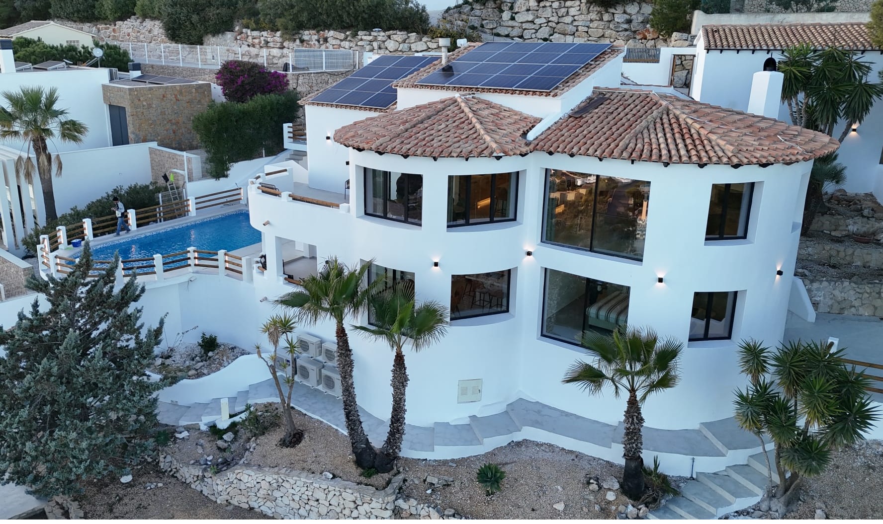 Außenansicht einer weißen, mehrstöckigen Villa mit geschwungenen Formen, Terrakotta-Ziegeldach mit Solarpaneelen und privatem Poolbereich mit Palmen.