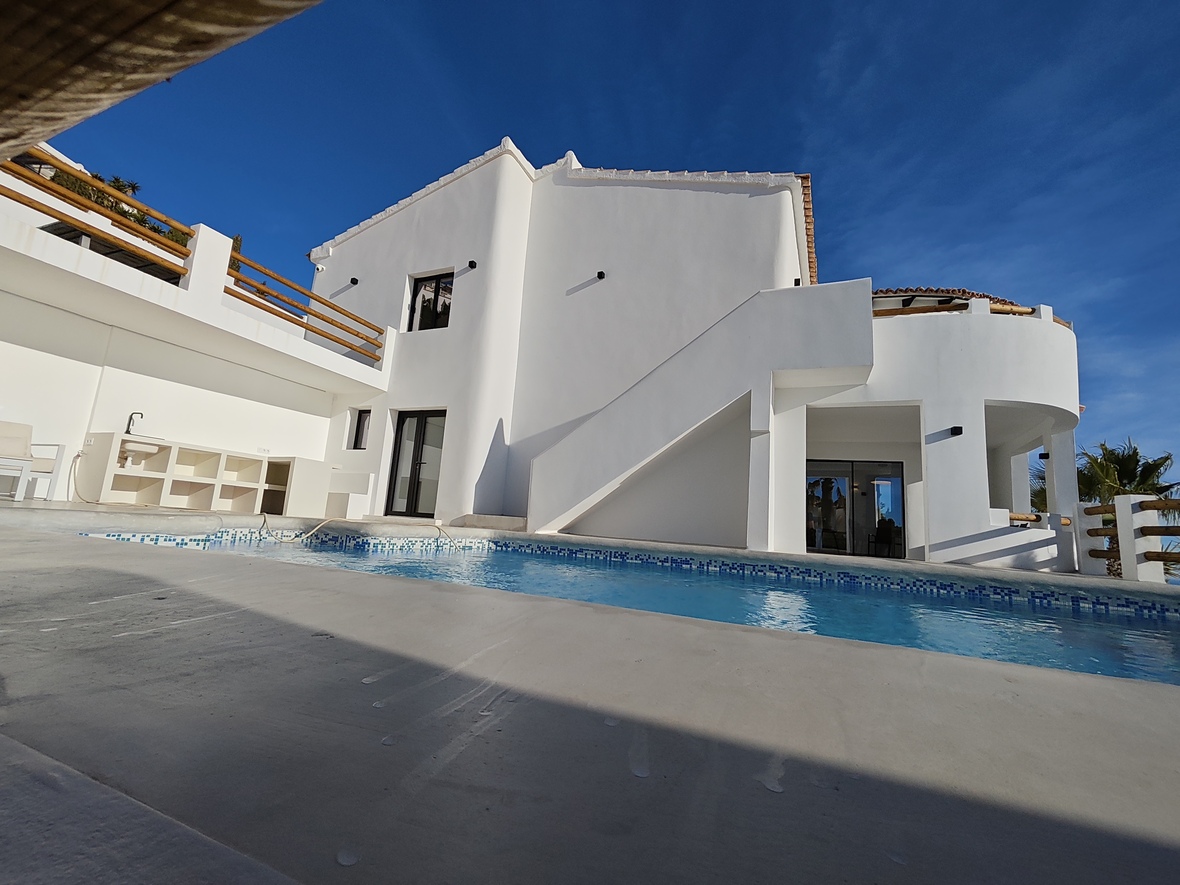Außenansicht einer weißen Villa mit blau gefliestem Pool, integrierter Außenküche und Holzgeländern auf mehreren Ebenen.