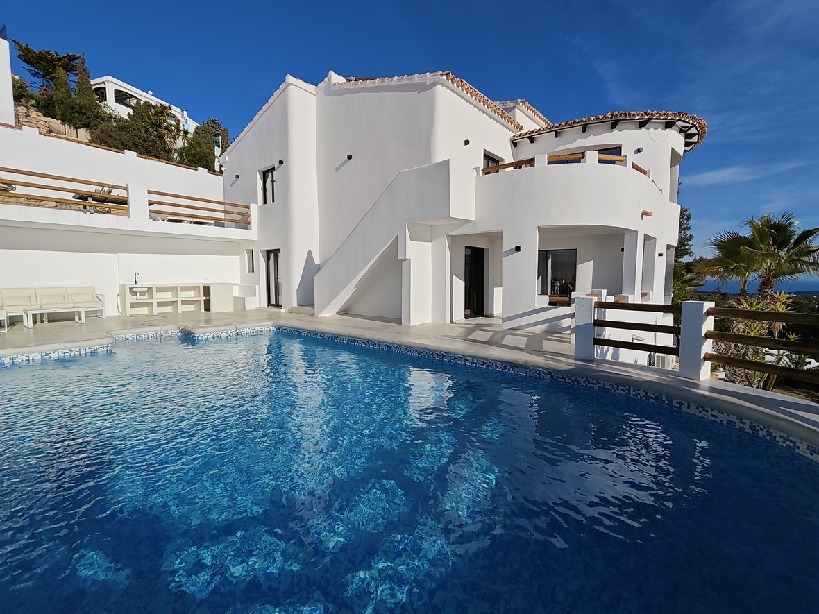 Außenansicht einer weißen Villa mit geschwungenem Pool, blauen Mosaikfliesen und Terrassen mit Geländern aus Naturholz.