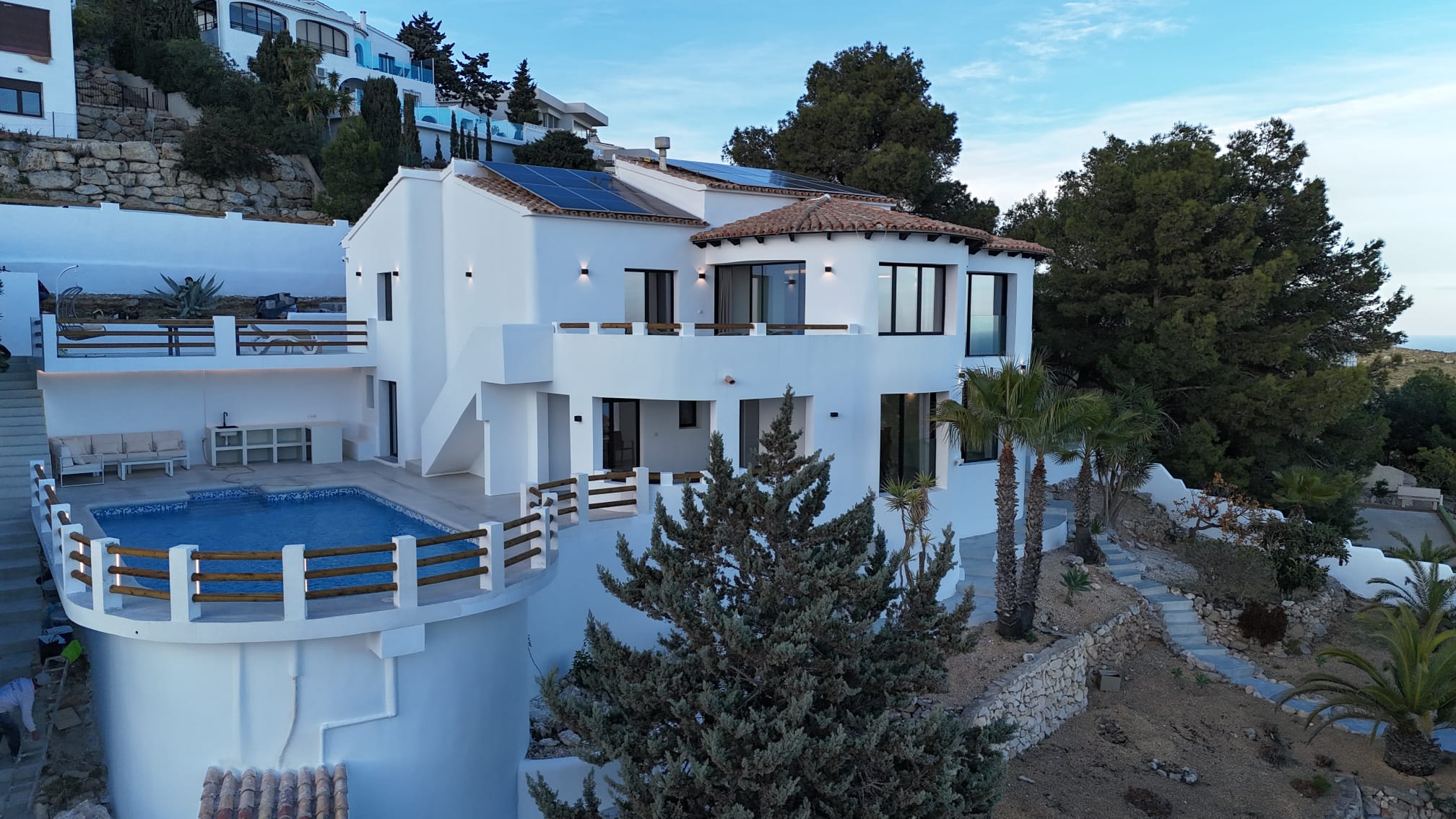 Außenansicht einer weißen Villa mit geschwungenem Pool, Solarmodulen auf dem Dach und terrassierten Steingärten mit mediterraner Vegetation.