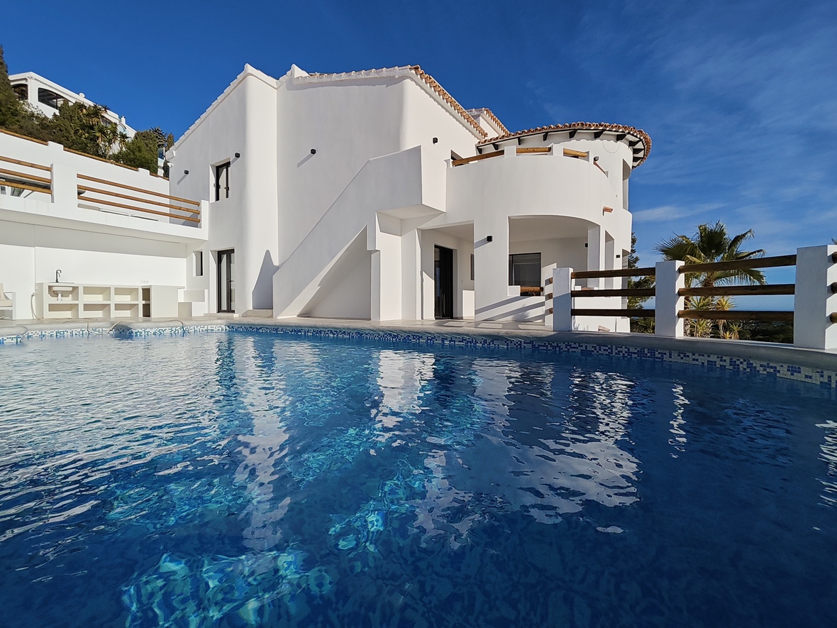Außenansicht einer weißen Villa mit geschwungenen Terrassen, Holzgeländern und einem blau gefliesten Pool, der die mediterrane Architektur spiegelt.