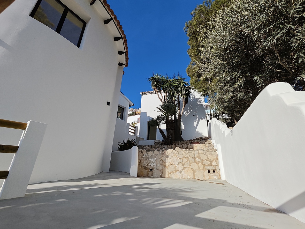 Außenansicht mit geschwungenen weißen Wänden, einer Natursteinmauer und gepflastertem Boden. Mediterrane Bepflanzung mit Palmen und Bäumen unter strahlend blauem Himmel.