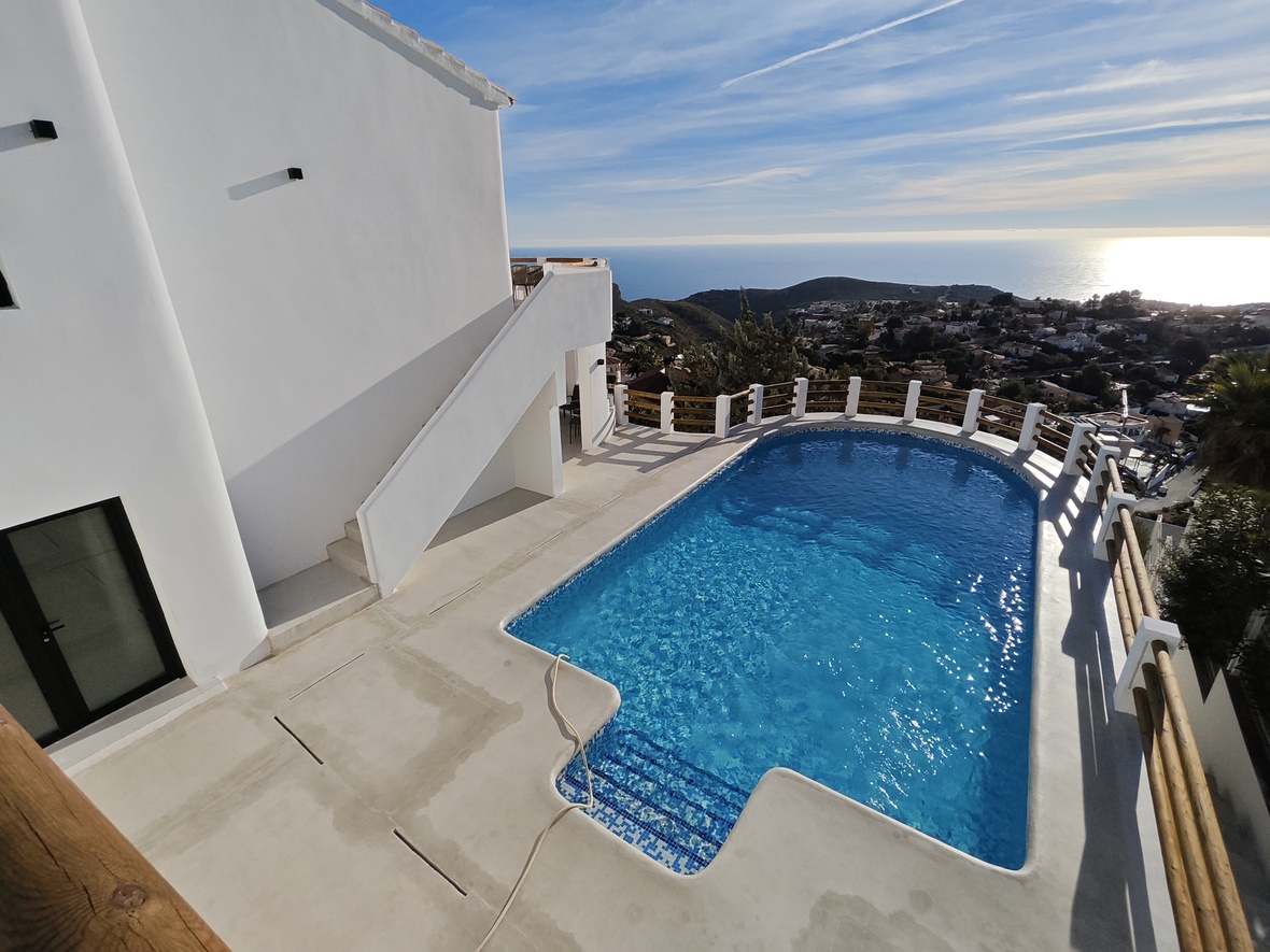 Außenpool mit blauen Mosaikfliesen und weißer Terrasse, eingefasst von Holzgeländern mit weitem Blick über das Mittelmeer und die Küstenlandschaft.