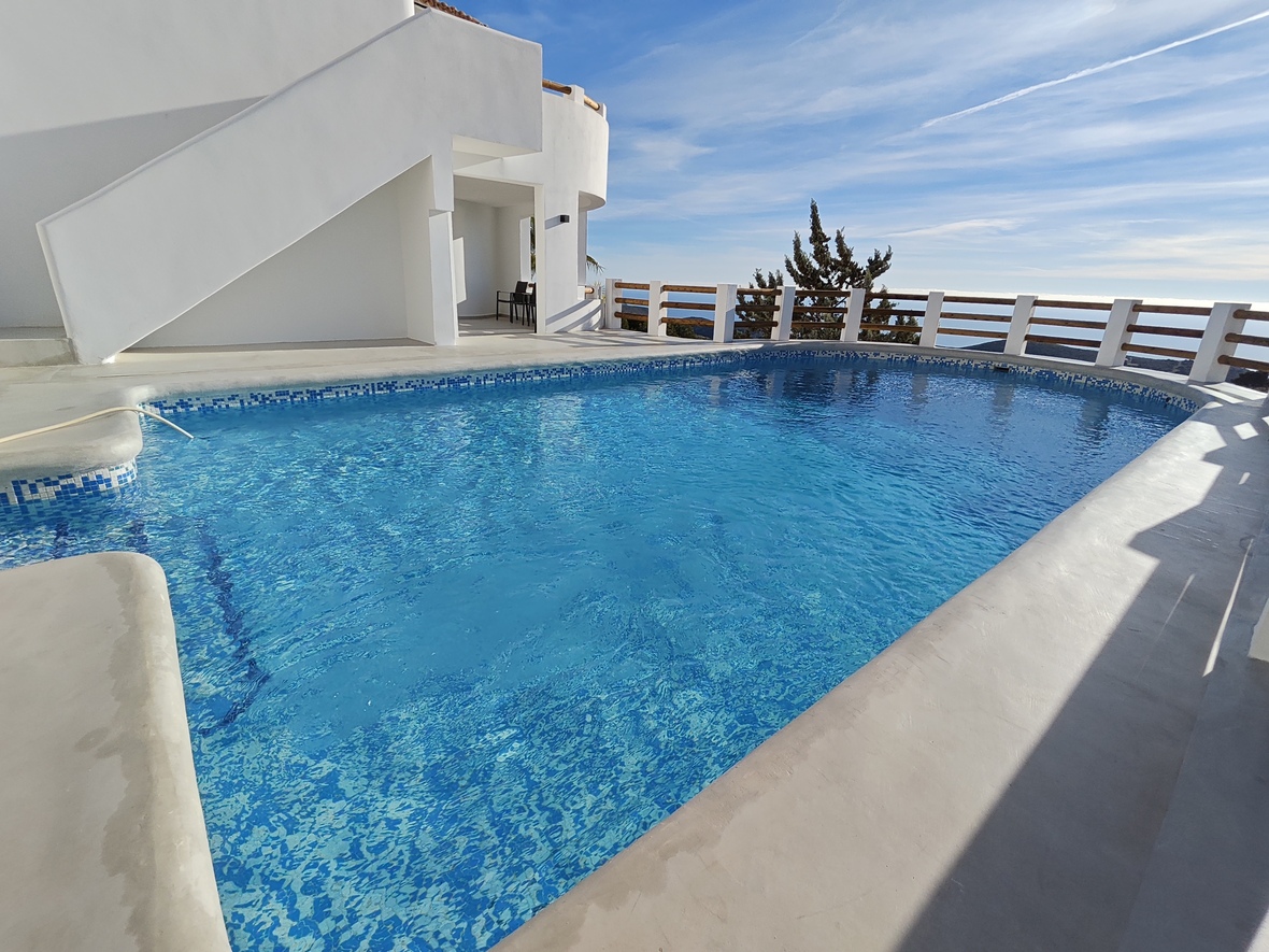 Außenpool mit blauen Mosaikfliesen, weißer Beton-Terrasse und Holzgeländer mit weitem Panoramablick über das Meer.